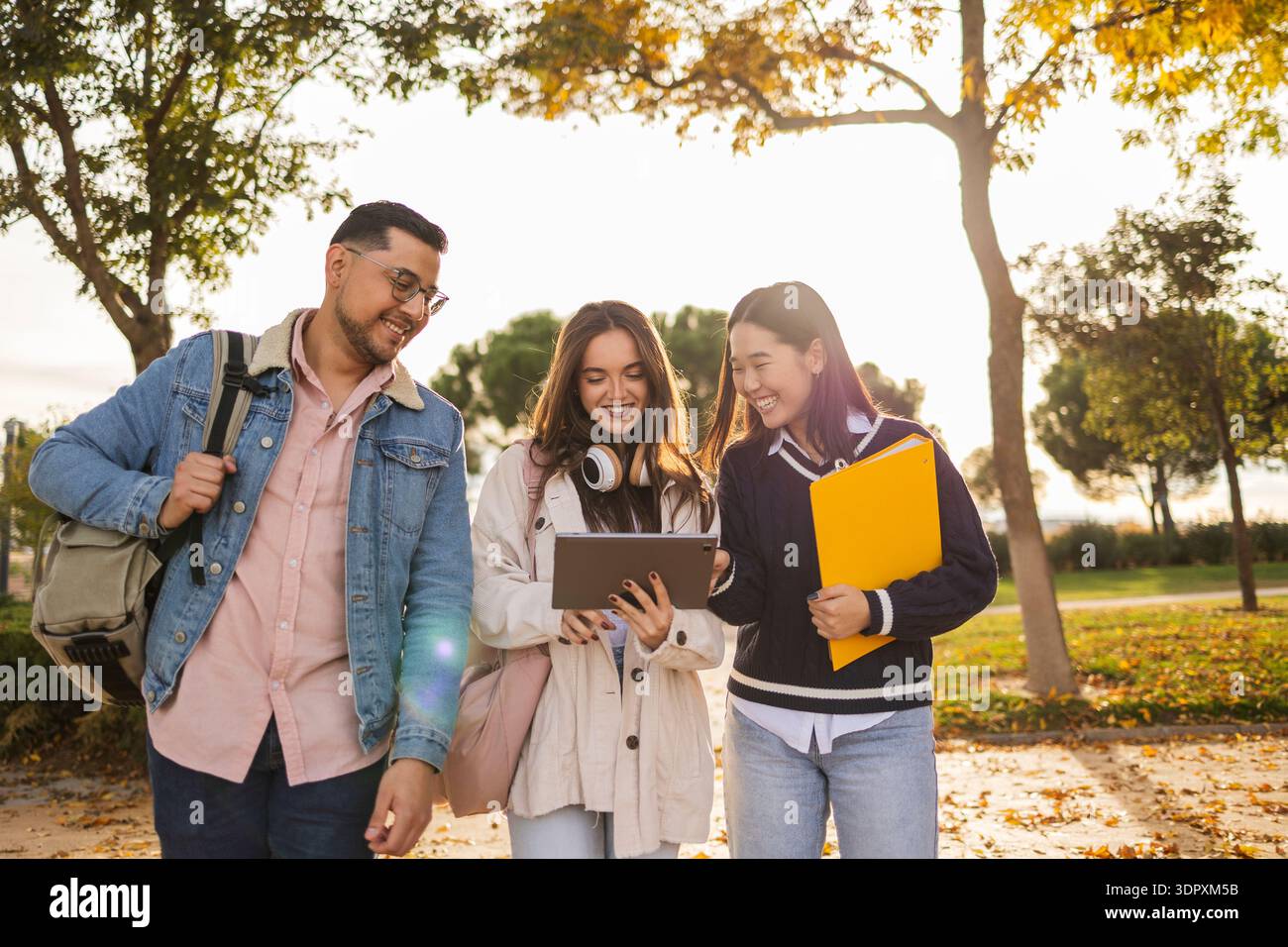 Un gruppo di tre diversi studenti universitari cammina insieme su un pittoresco percorso del campus, coinvolgendosi con un tablet e portando con sé libri, godendosi un au vivace Foto Stock