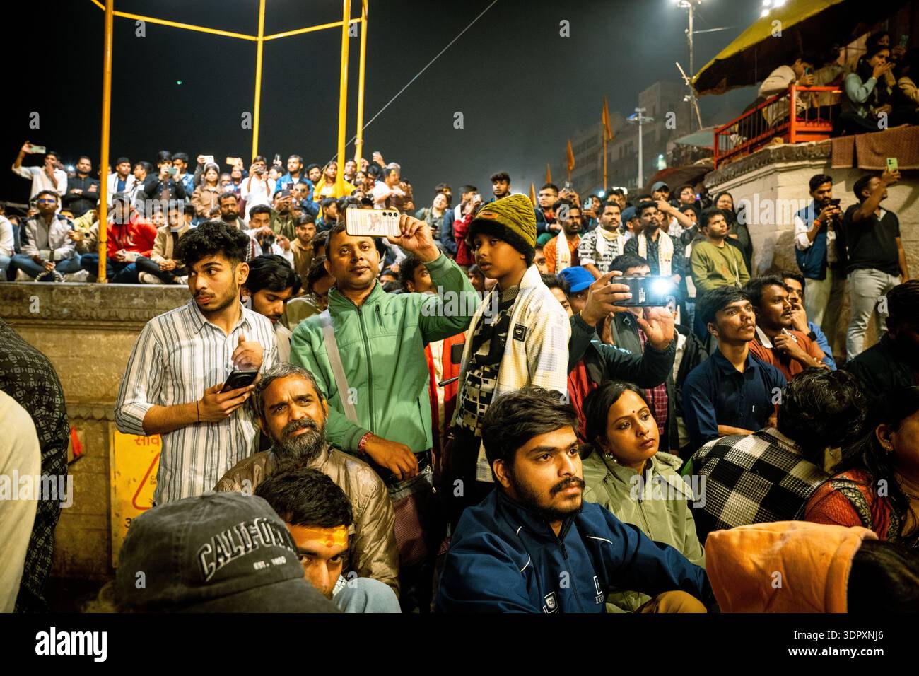 Una folla diversificata in India guarda un evento di notte, molti catturano l'attimo con smartphone e fotocamere. Foto Stock