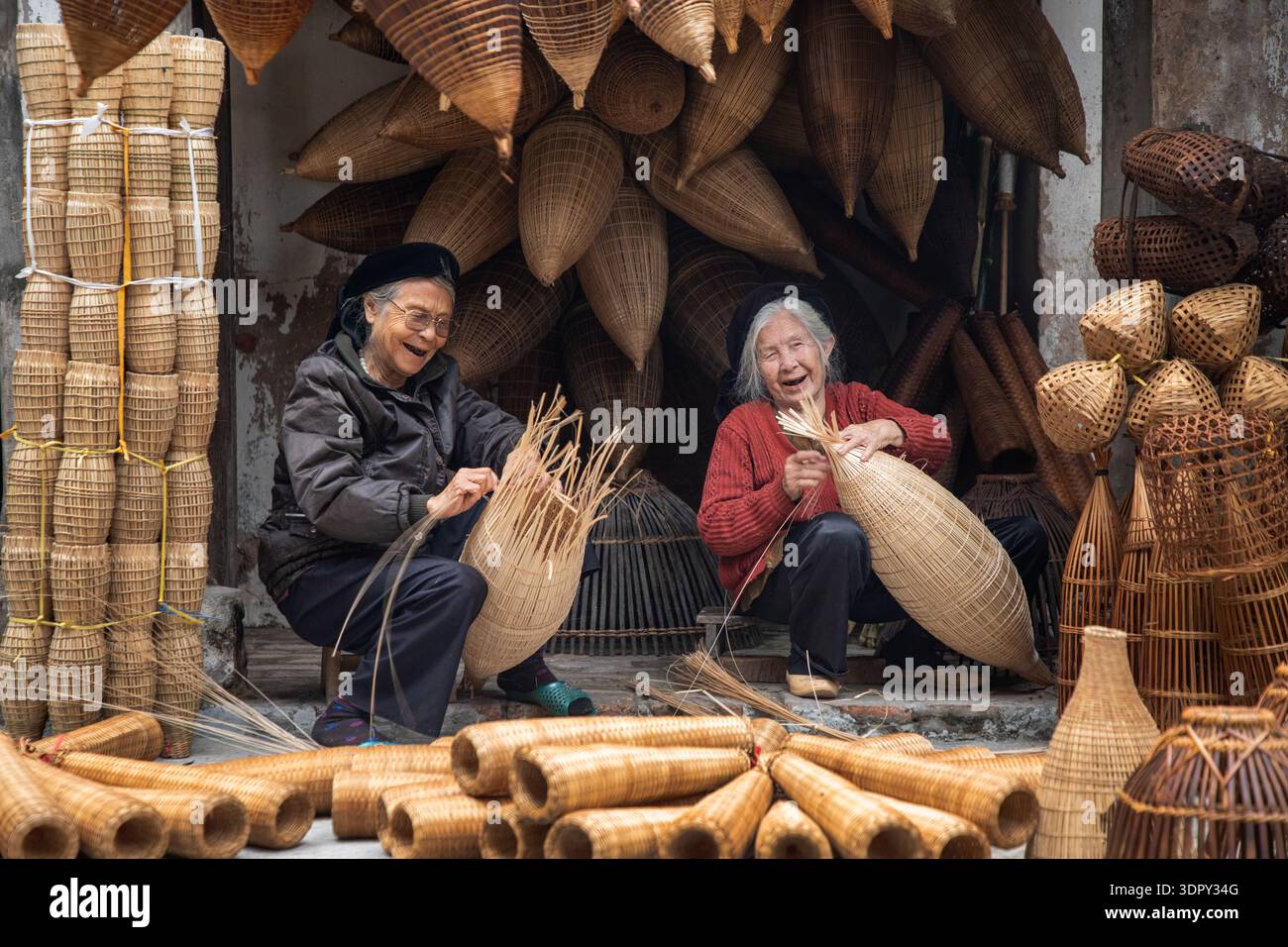 Hưng Yên, Vietnam - 15 dicembre 2019: Vista di due donne che ridono mentre tessono intricate trappole di pesci di bambù, circondate da una calda e terrosa tavolozza di ettari Foto Stock
