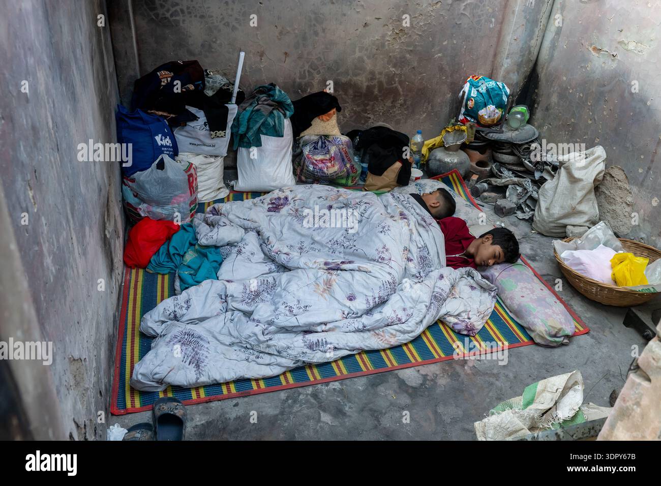 Dacca, Bangladesh - 28 novembre 2025: Vista di una camera poco illuminata dove due personaggi dormono sonni tranquilli su un tappeto a righe, circondato da borse e belon personale Foto Stock