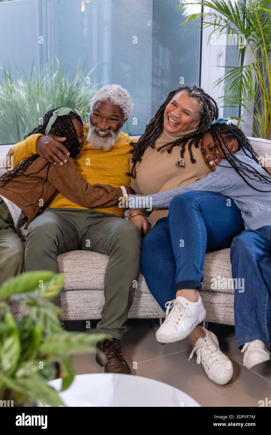 Famiglia afroamericana che si coccola sul divano di casa, senior che abbraccia due bambine al tavolino da caffè Foto Stock