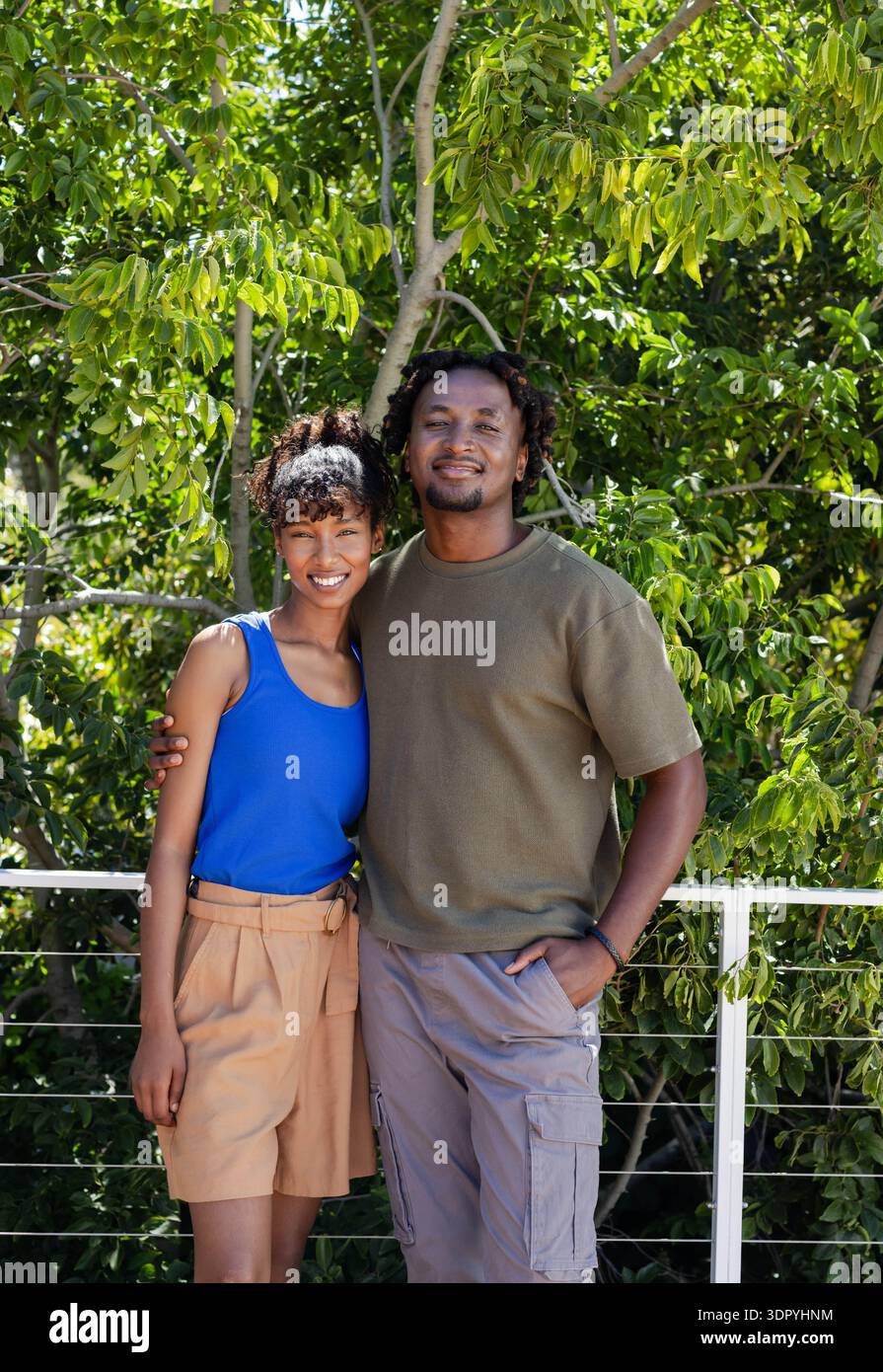 Coppia afroamericana sorridente sul balcone, indossa canotta e pantaloni cargo, reggendo ringhiere a cavo Foto Stock