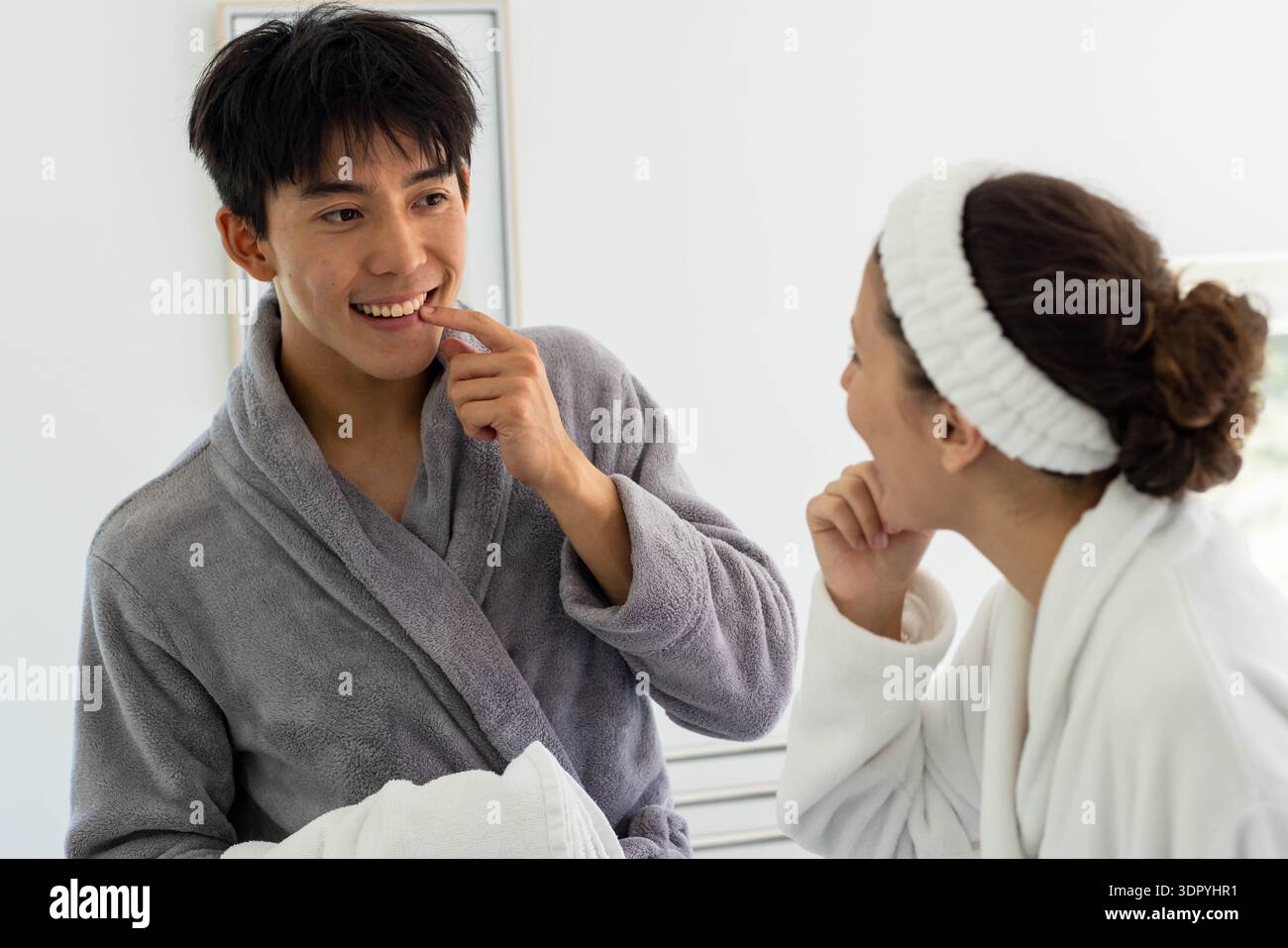 Diverse coppie in accappatoi che si puliscono vicino allo specchio del bagno, un dente che tocca con un asciugamano e l'altro guarda Foto Stock