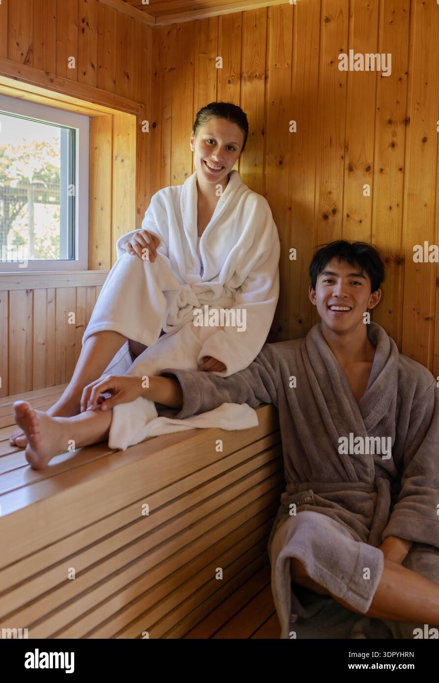 Coppia seduta su panche sauna in legno con accappatoi bianchi e grigi, sorridendo verso la macchina fotografica Foto Stock