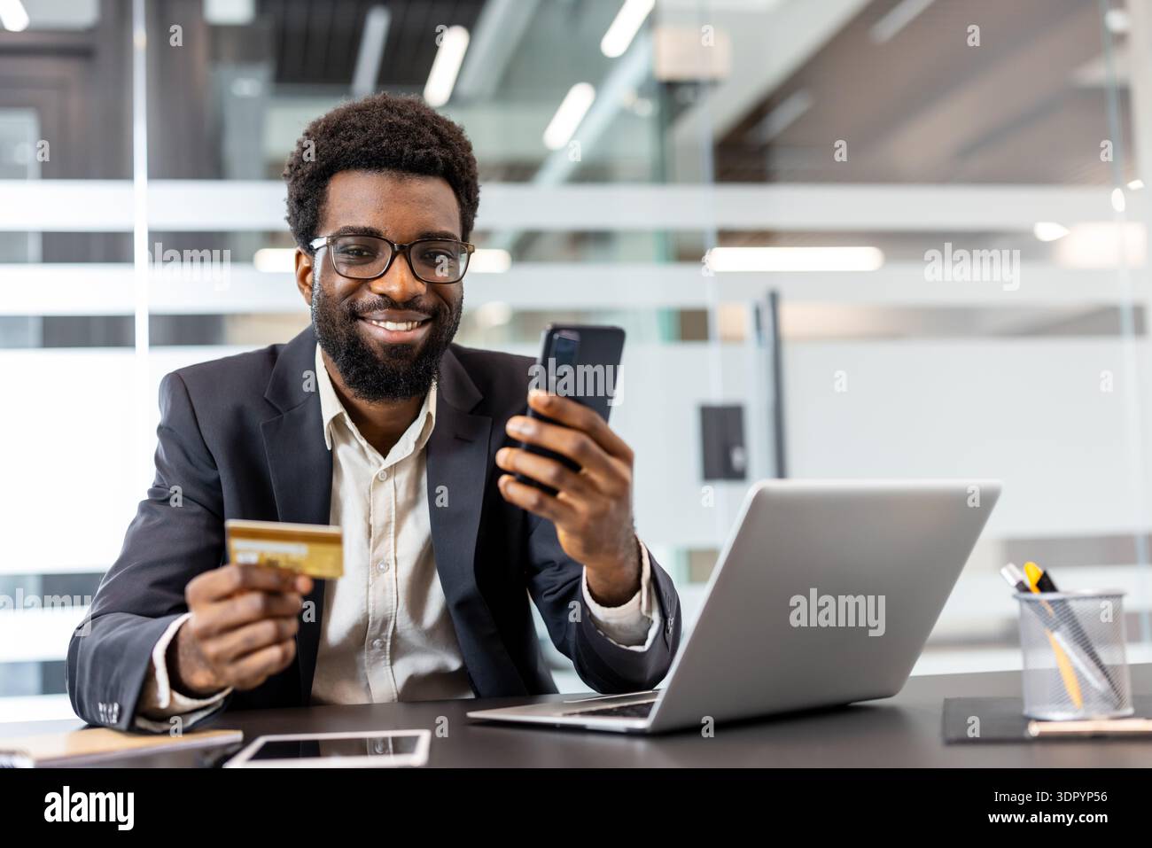 Un uomo sorridente completa un pagamento mobile sicuro con una carta di credito e uno smartphone dorati alla sua scrivania con un computer portatile, illustrando e-commerce, servizi bancari e transazioni digitali moderne Foto Stock