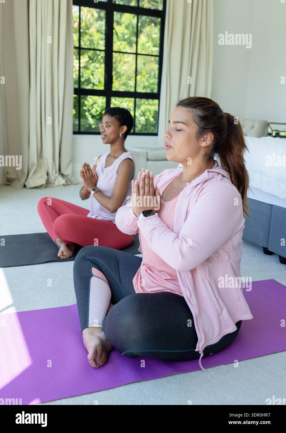 Due amiche in abbigliamento sportivo sedute su tappeti scuri viola che meditano in camera da letto con l'orologio Foto Stock
