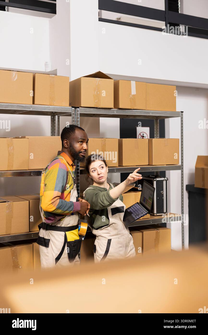 Due dipendenti in tute che supervisionano il controllo dell'inventario e il confezionamento in un piccolo deposito, preparando i prodotti per le spedizioni di ordini personalizzati. Servizi di e-commerce interni e spedizione accurata. Foto Stock