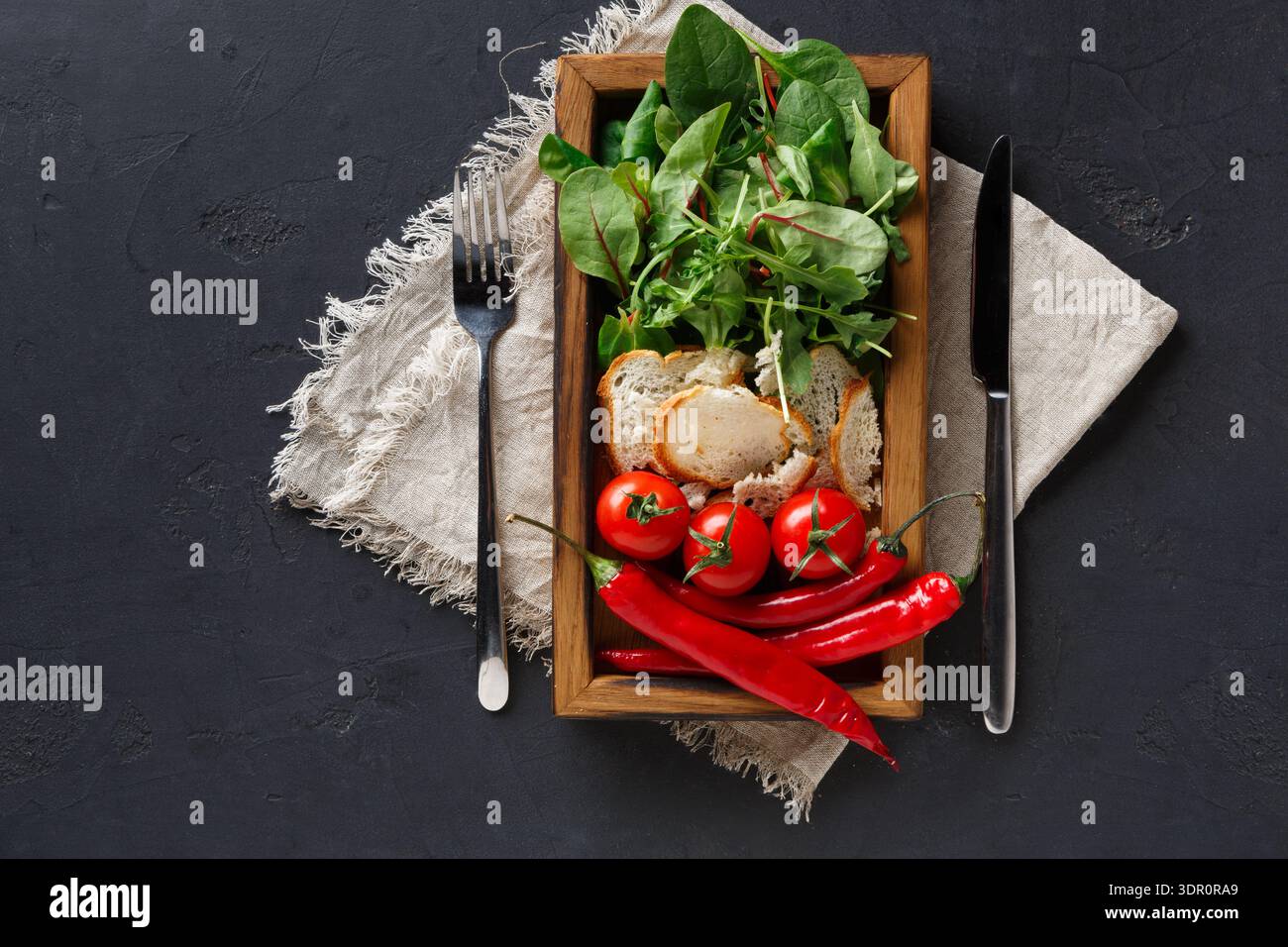 Verdure fresche e pane disposti su un piatto di legno con utensili su sfondo scuro Foto Stock