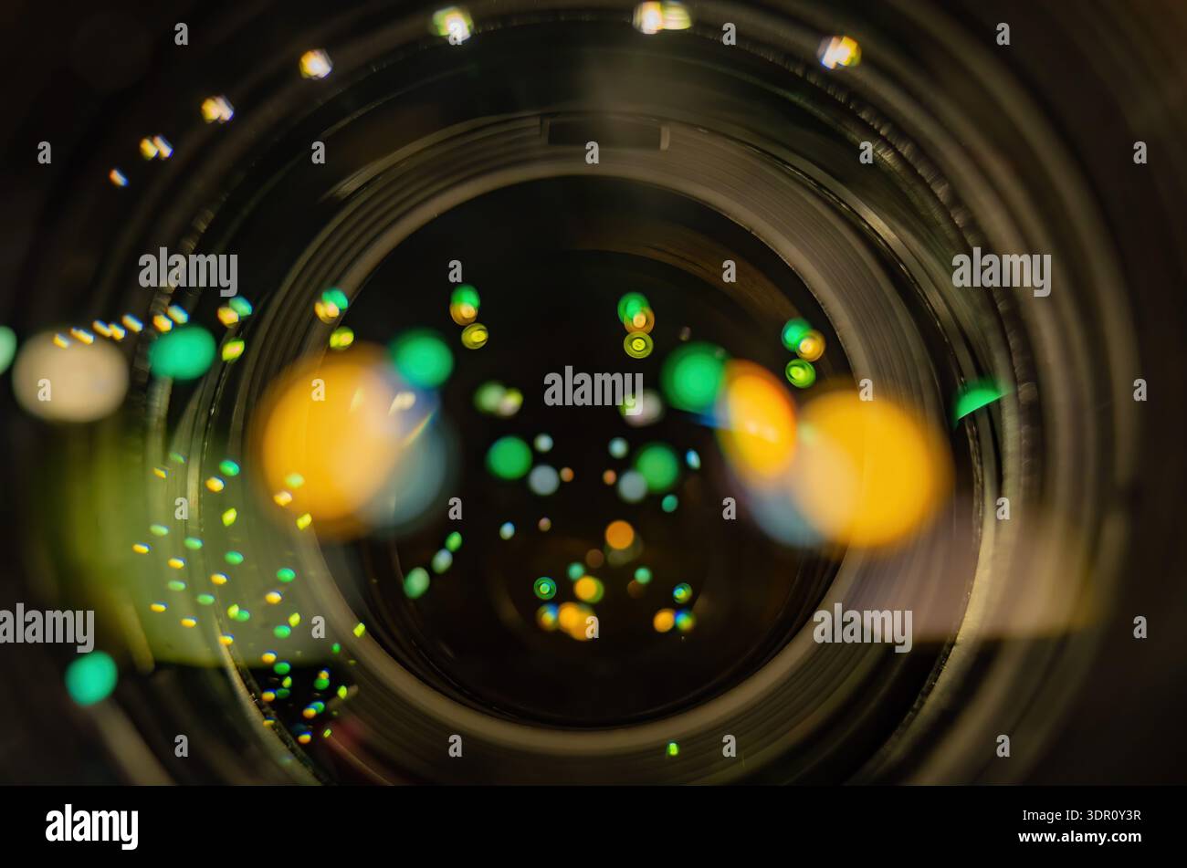 Le vivaci luci bokeh verdi e arancioni si illuminano attraverso l'apertura circolare dell'obiettivo di una fotocamera in un ambiente buio. Foto Stock