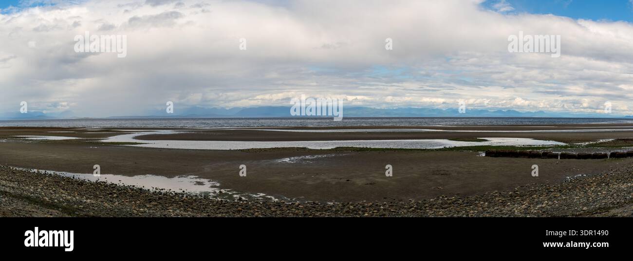 Un'ampia vista panoramica cattura una spiaggia rocciosa, montagne lontane e un cielo per lo più nuvoloso lungo la costa dell'isola di Vancouver. Foto Stock