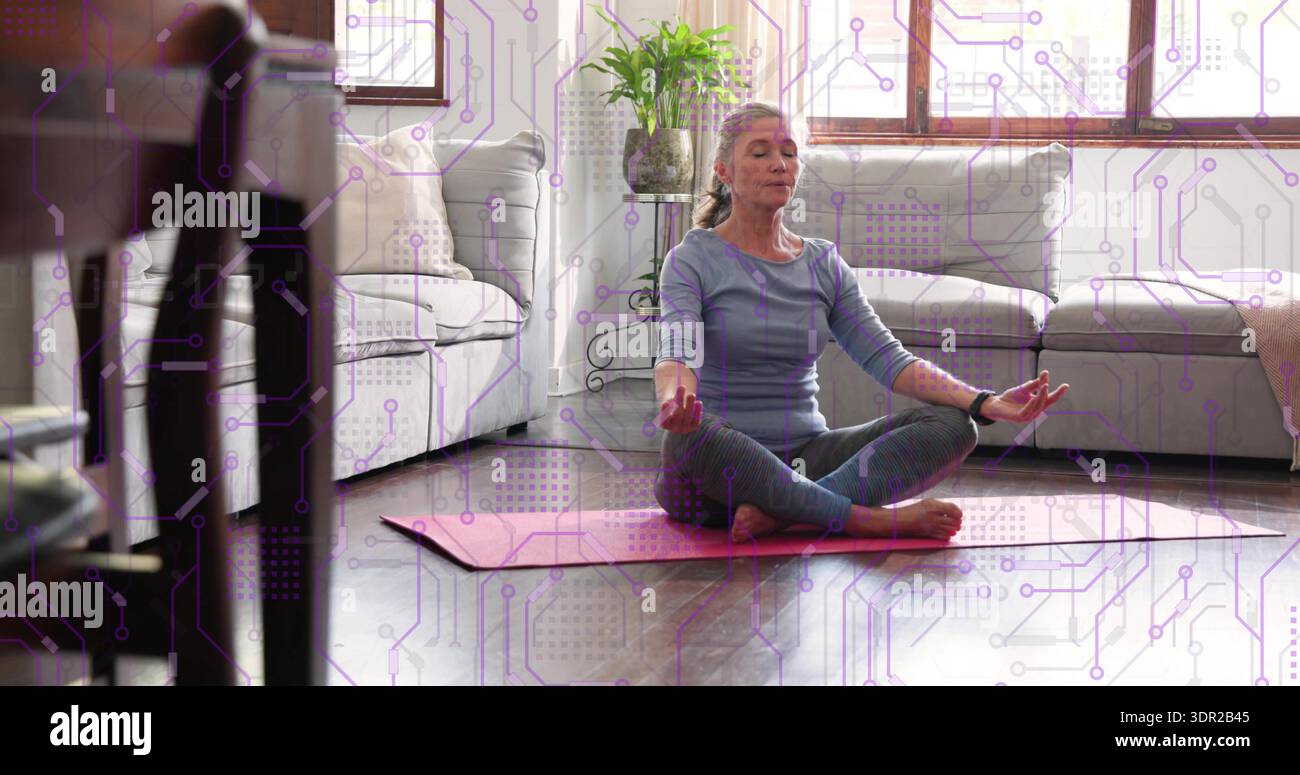 Donna matura seduta a meditare a gambe incrociate nel soggiorno di casa, indossando abiti da yoga su un tappetino rosa Foto Stock