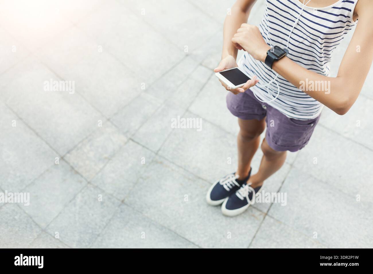 La persona che indossa una camicia a righe scambia lo smartwatch mentre tiene il telefono in posizione esterna alla luce del giorno Foto Stock