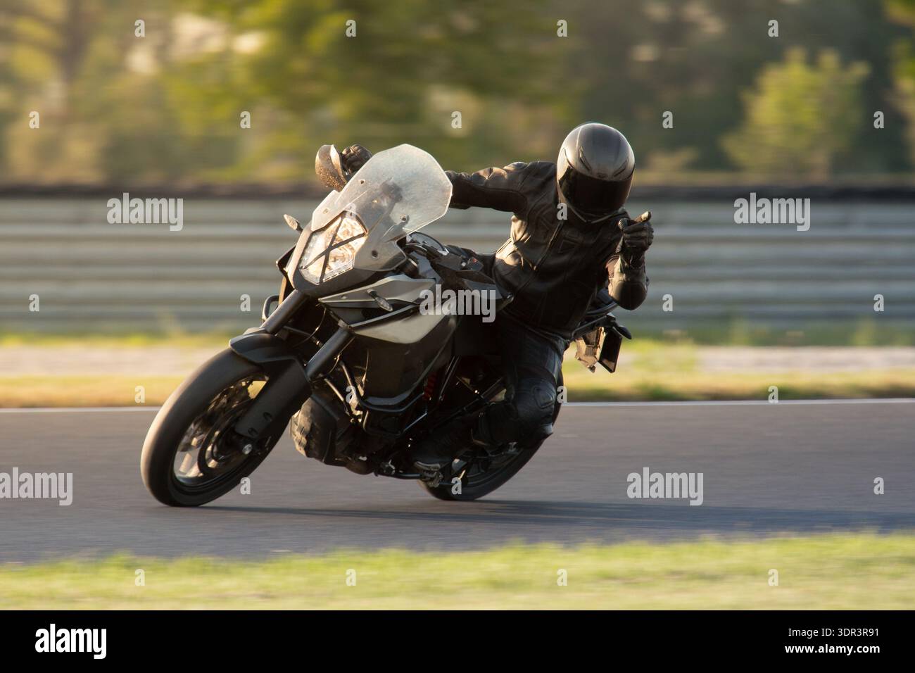 Un motociclista con l'equipaggiamento protettivo nero che si appoggia a una motocicletta sportiva grigia in una curva su un circuito durante una giornata di pista al tramonto. Foto Stock