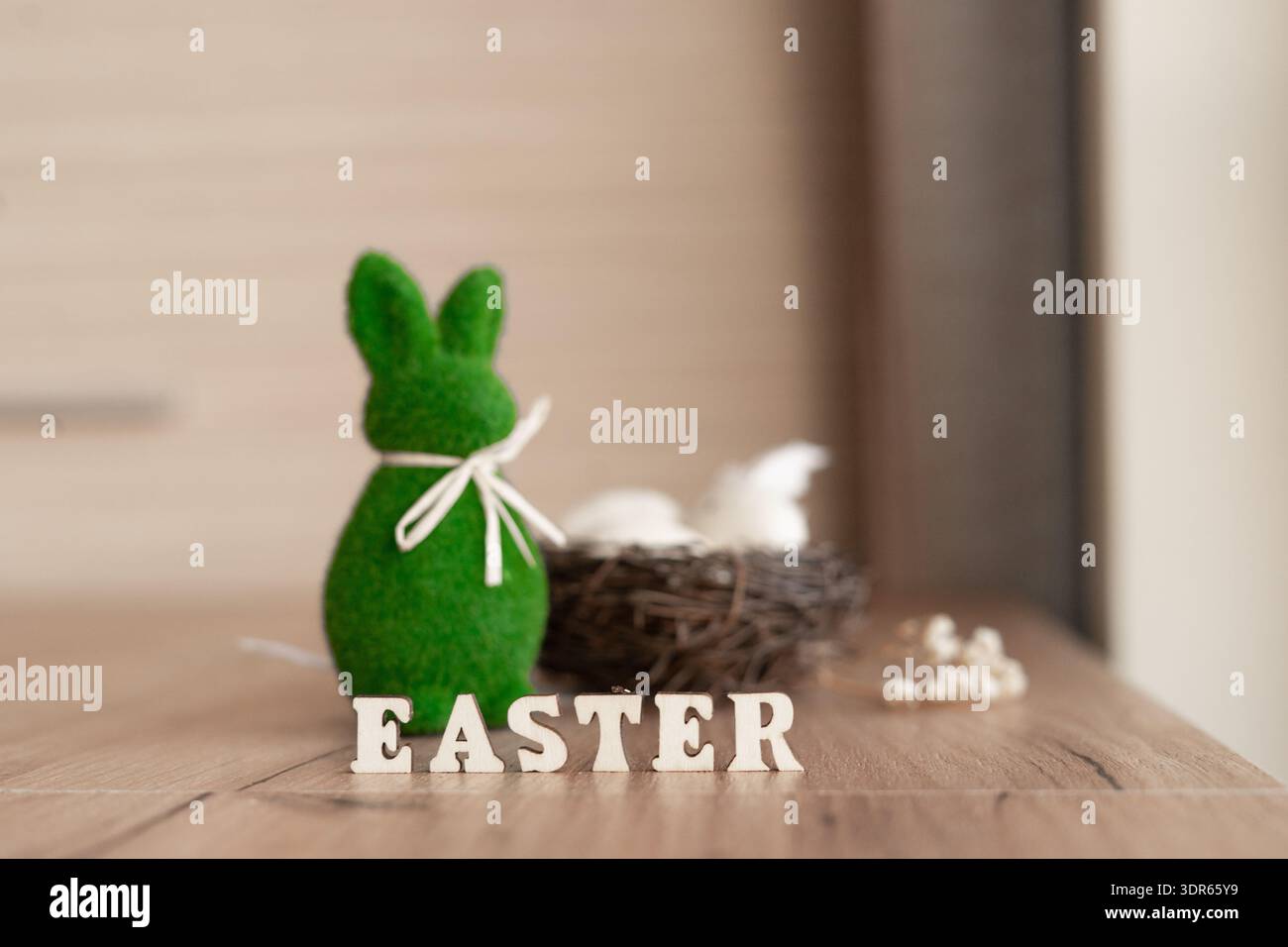 Natura morta pasquale con coniglietto verde e nido su tavola rustica. Verde feltro coniglio con nastro bianco accanto al nido di vimini e uova, lettere di legno ortografiche Foto Stock