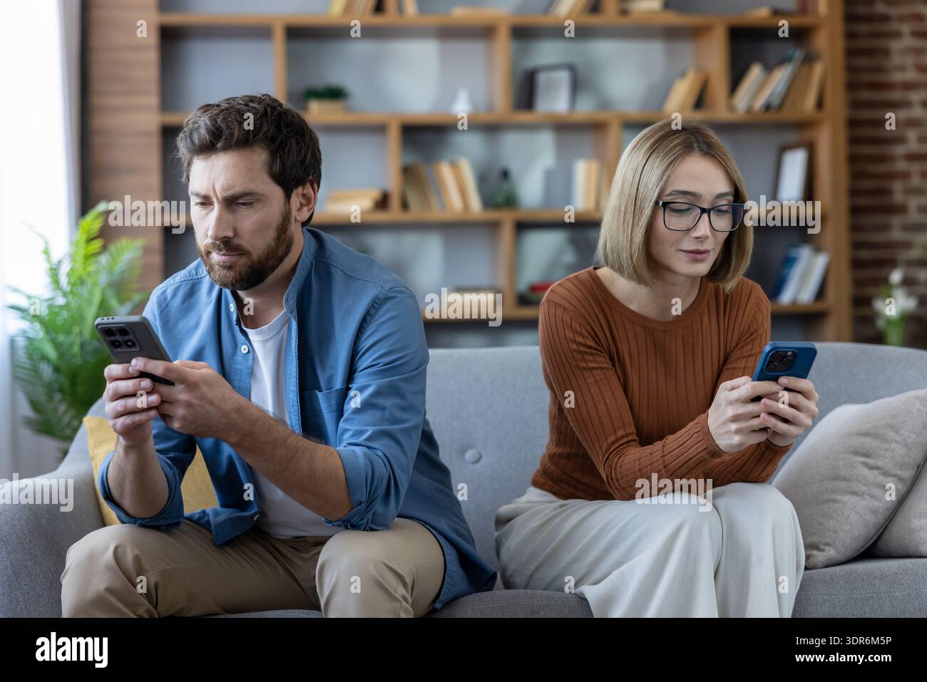Coppia seduta su schermi rivolti al divano, ciascuno assorbito dal proprio smartphone mentre scena domestica emotivamente lontana di disconnessione digitale, scorrimento Foto Stock