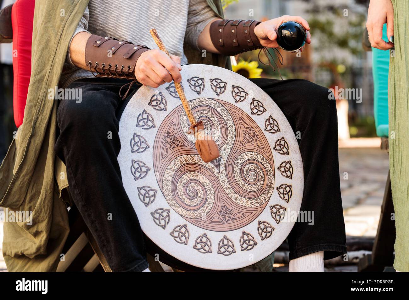 Una persona con un abbigliamento in stile medievale suona un grande tamburo decorato mentre tiene in mano una palla di cristallo nero, suggerendo un contesto spirituale o cerimoniale Foto Stock