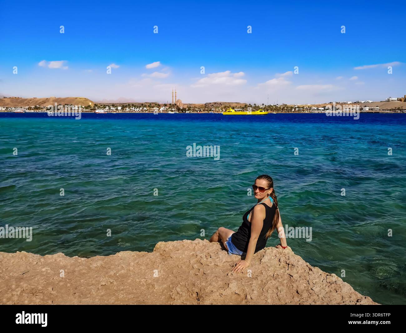 Il turista europeo siede sul bordo di una scogliera sopra le acque turchesi-azzurre del Mar Rosso a Sharm El Sheikh (Egitto). Giovane donna in abiti estivi Foto Stock