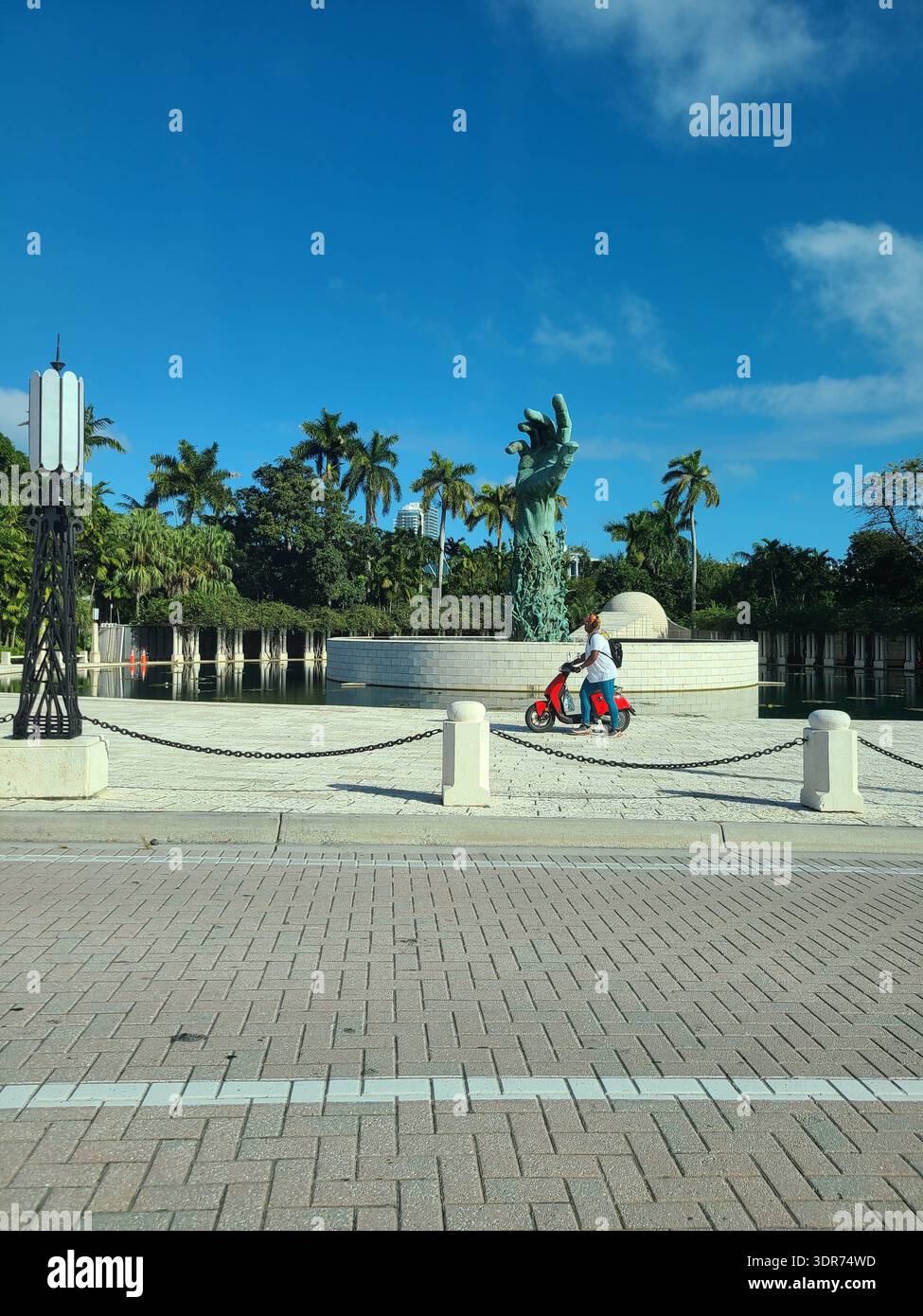SOLO USO EDITORIALE - MIAMI FLORIDA: La potente "scultura dell'amore e dell'angoscia" al Memoriale dell'Olocausto di Miami Beach, Florida. Una grande mano in bronzo raggiunge il cielo blu luminoso da una piazza circolare in pietra circondata da una piscina riflettente e palme. In primo piano, una persona su uno scooter rosso passa lungo la strada lastricata di mattoni sotto un cielo tropicale limpido e soleggiato. Foto Stock