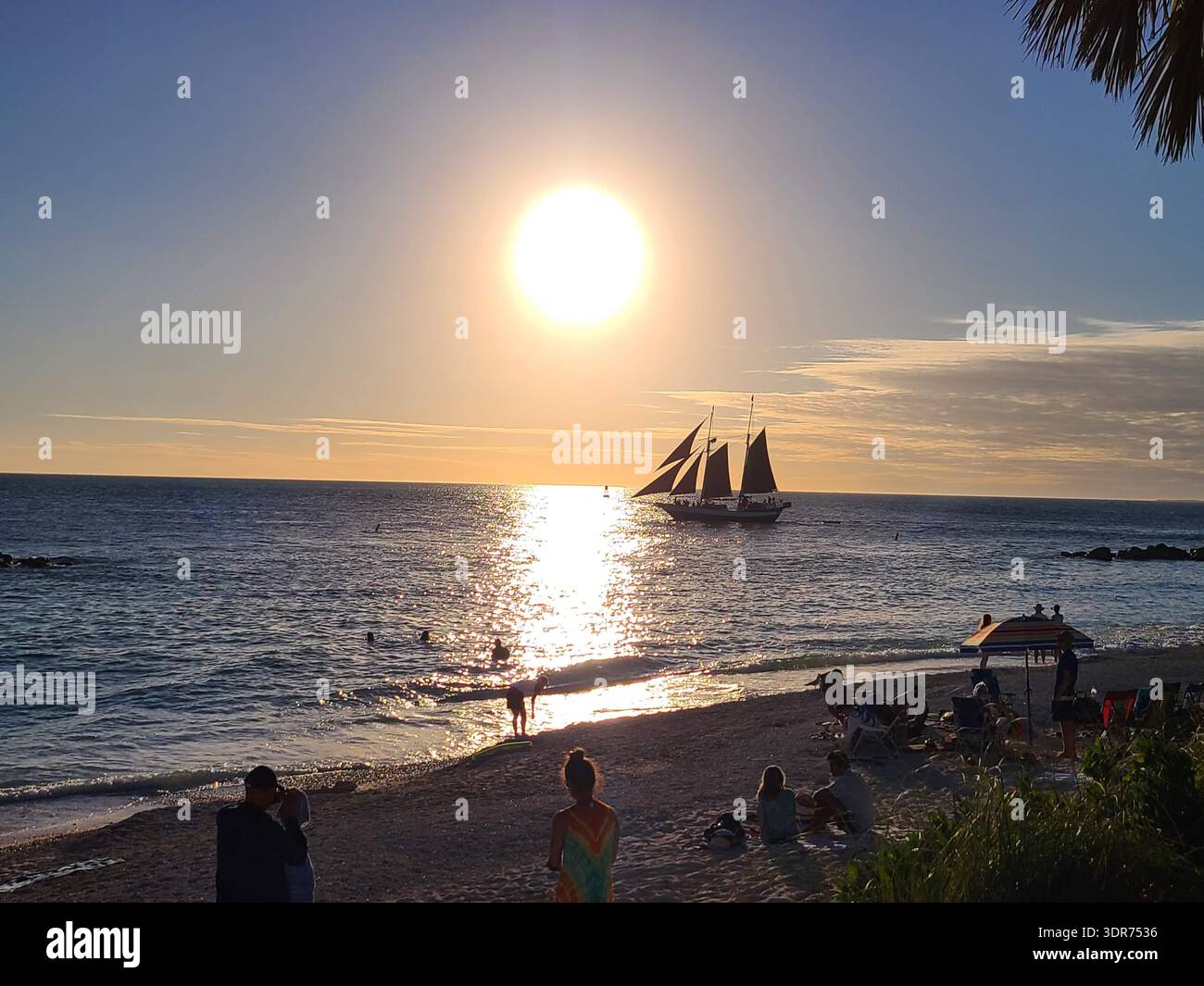 SOLO PER USO EDITORIALE - KEY WEST FLORIDA: Sagoma di una nave a vela e turisti a Fort Zachary Taylor Historic State Park Beach, Key West. Un paesaggio da tramonto grandangolare caratterizzato da un'astronave sagomata contro un sole luminoso sul Golfo del Messico. In primo piano, i turisti si rilassano sulla riva di una spiaggia tropicale a Key West, Florida. La scena cattura l'iconica celebrazione quotidiana del tramonto e l'atmosfera tropicale della città più meridionale degli Stati Uniti continentali - Immagine stock catturata con smartphone