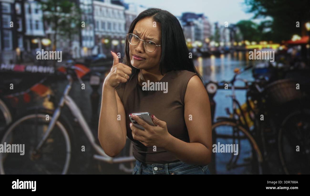 Donna che tiene in mano lo smartphone, controlla i messaggi con un gesto premuroso del pollice e indossa gli occhiali in studio; curiosità scettica. Foto Stock