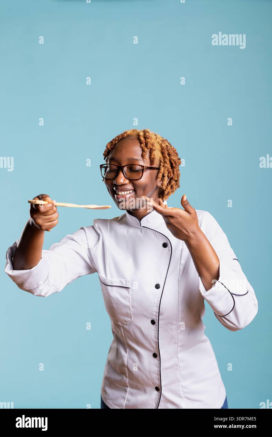 Allegra chef donna che assaggia cibo in modo uniforme utilizzando un cucchiaio di legno su sfondo blu. Cuoco professionista che esprime soddisfazione e creatività, mostrando passione ed eccellenza per le arti culinarie Foto Stock