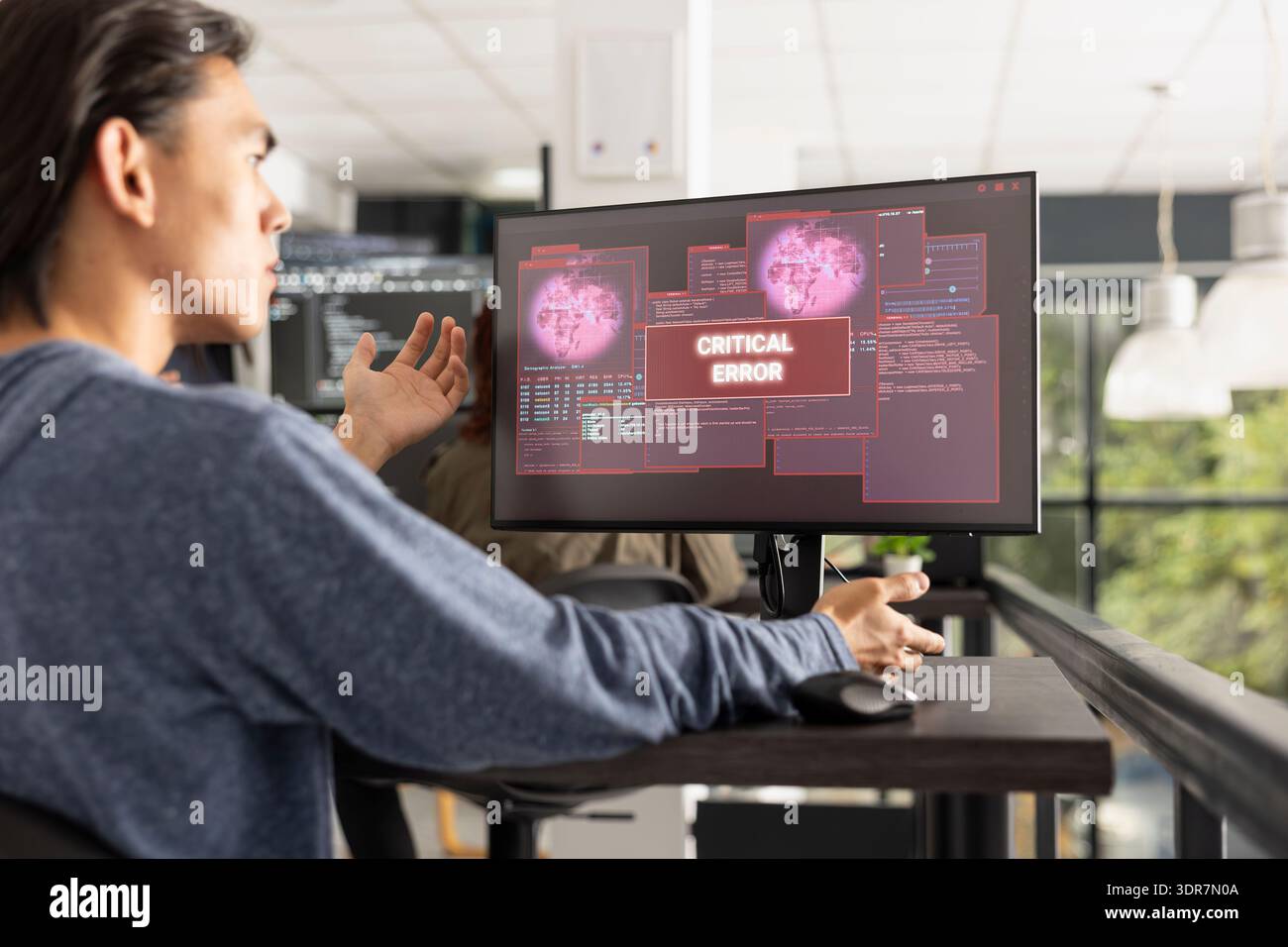 Il programmatore asiatico esamina il monitor del computer mostrando un avviso di errore critico. Il programmatore focalizzato risponde con preoccupazione, sottolineando le problematiche di debug, codifica e risoluzione dei problemi software. Foto Stock