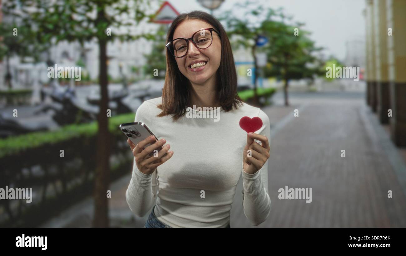 Donna in occhiali con un piccolo cuore rosso e sorridente mentre legge un messaggio sul suo smartphone in jeans casual su una strada della città costeggiata da alberi; ro Foto Stock