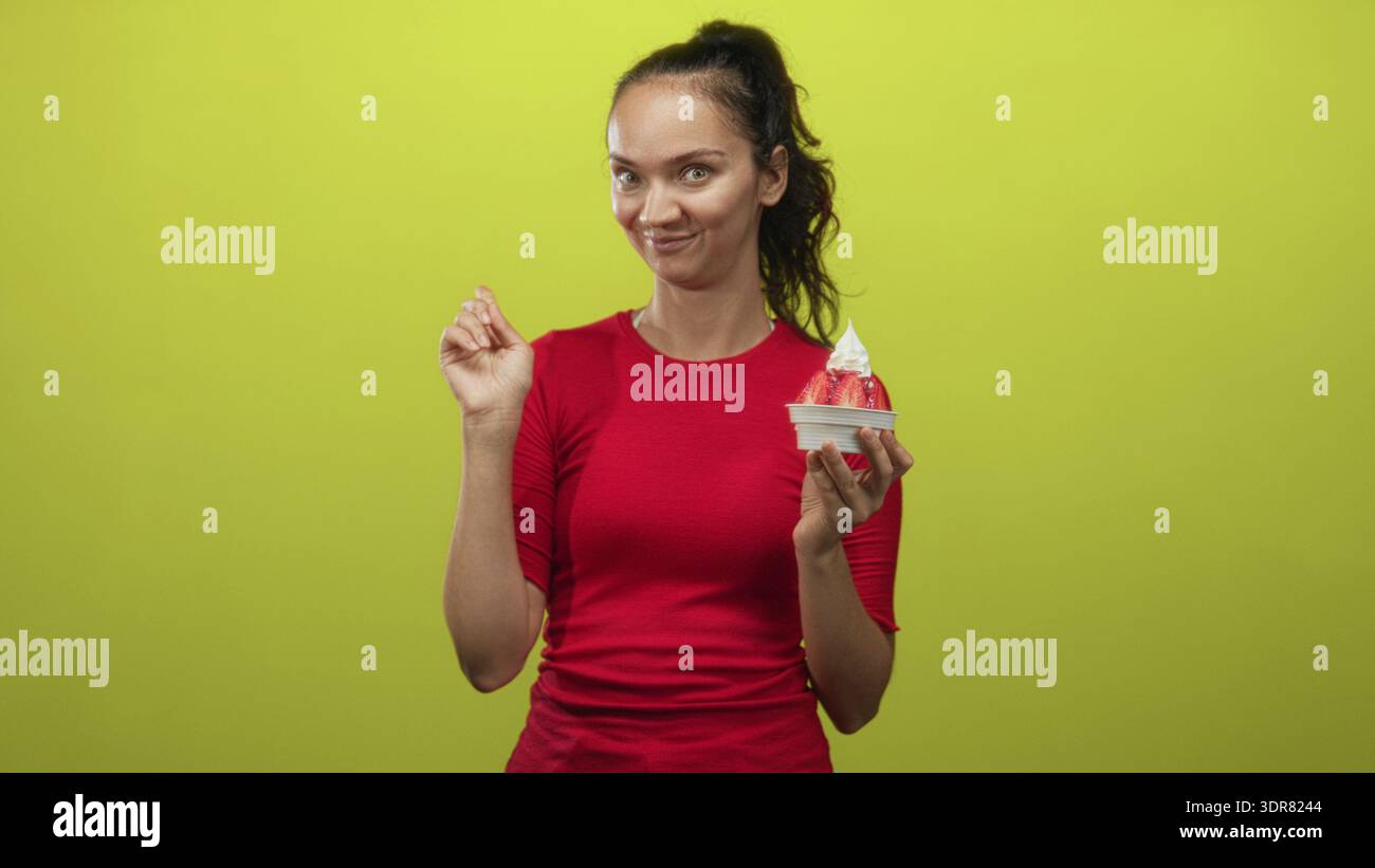 Donna che tiene in mano una tazza di gelato alle fragole con fragole a fette, punta il dito con la mano mentre sorride sullo sfondo verde dello studio; playf Foto Stock