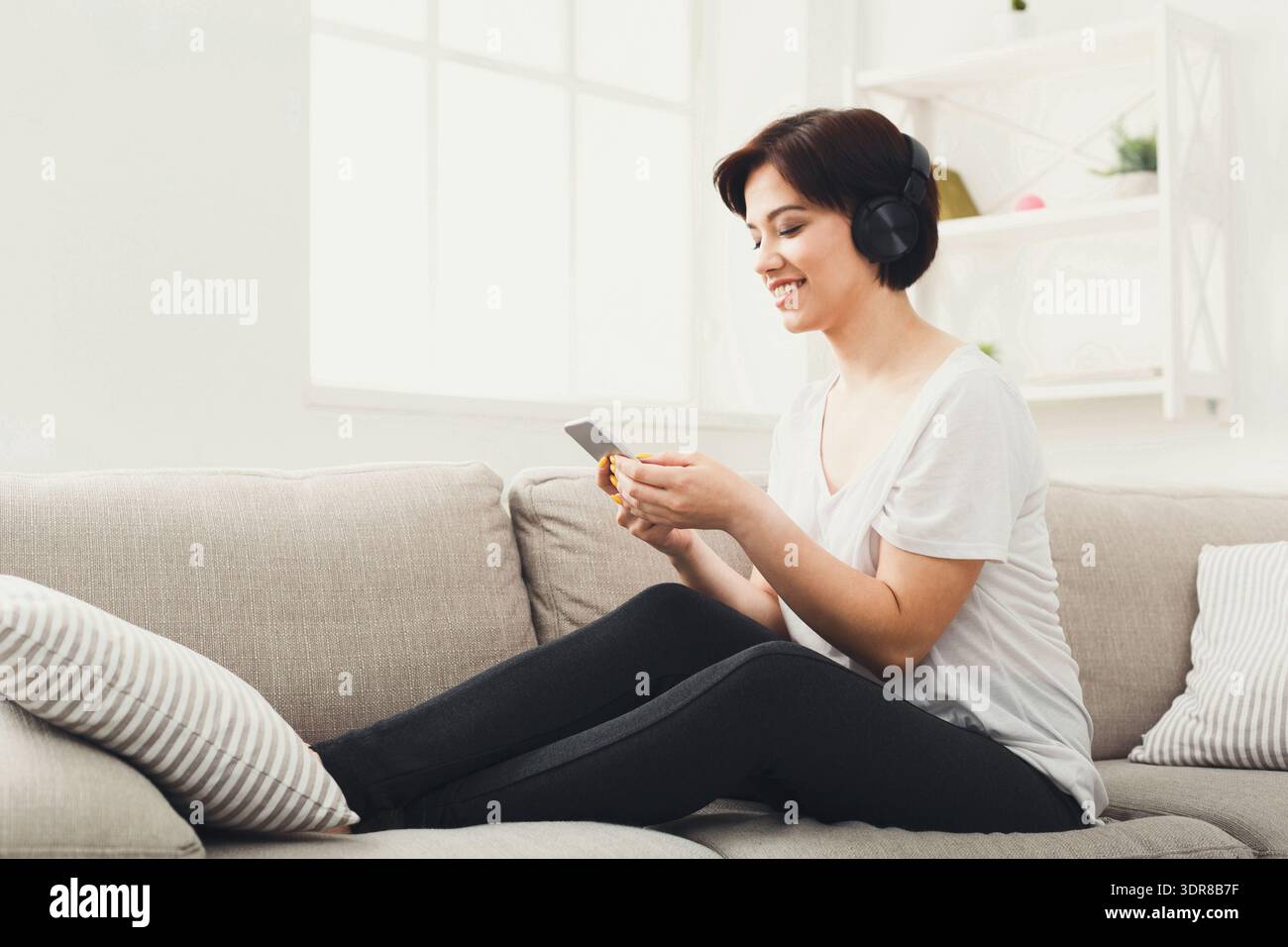 Donna seduta sul divano che ascolta musica sul telefono mentre indossa le cuffie Foto Stock
