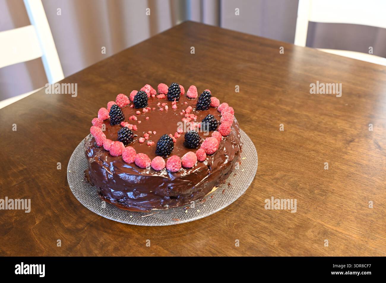 Torta al cioccolato ricoperta di lamponi freschi e ganache lucide su banco di vetro, dessert fatto in casa su un tavolo di legno con luce naturale soffusa. Foto Stock