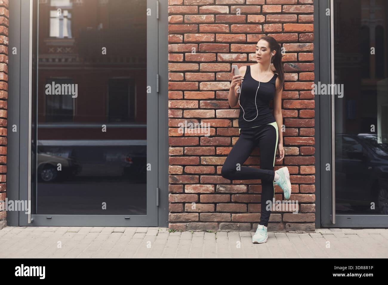 Donna in abiti da ginnastica si appoggia contro il muro di mattoni mentre ascolta musica sullo smartphone Foto Stock