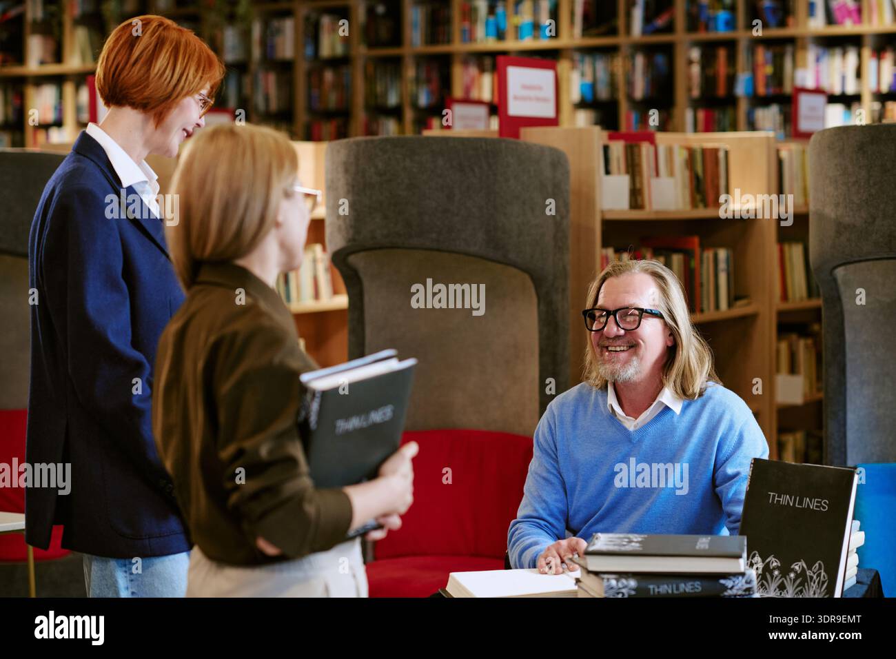 Uomo di mezza età caucasico che firma libri per due donne caucasiche in biblioteca Foto Stock