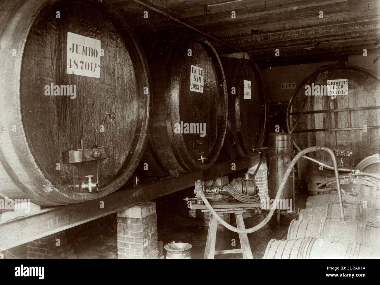 La fotografia mostra grandi barili di legno collegati a una pompa elettrica, con nomi come Jumbo, zio Sam, Bacchus e Groote Beer. I barili hanno capacità che vanno da 4.870 a 7.040 litri. L'immagine illustra lo stoccaggio industriale dei primi anni del XX secolo, i sistemi di gestione dei fluidi, la cura artigianale nella costruzione di fusti e l'infrastruttura meccanica per i liquidi in una fabbrica nei Paesi Bassi nel 1918. Foto Stock