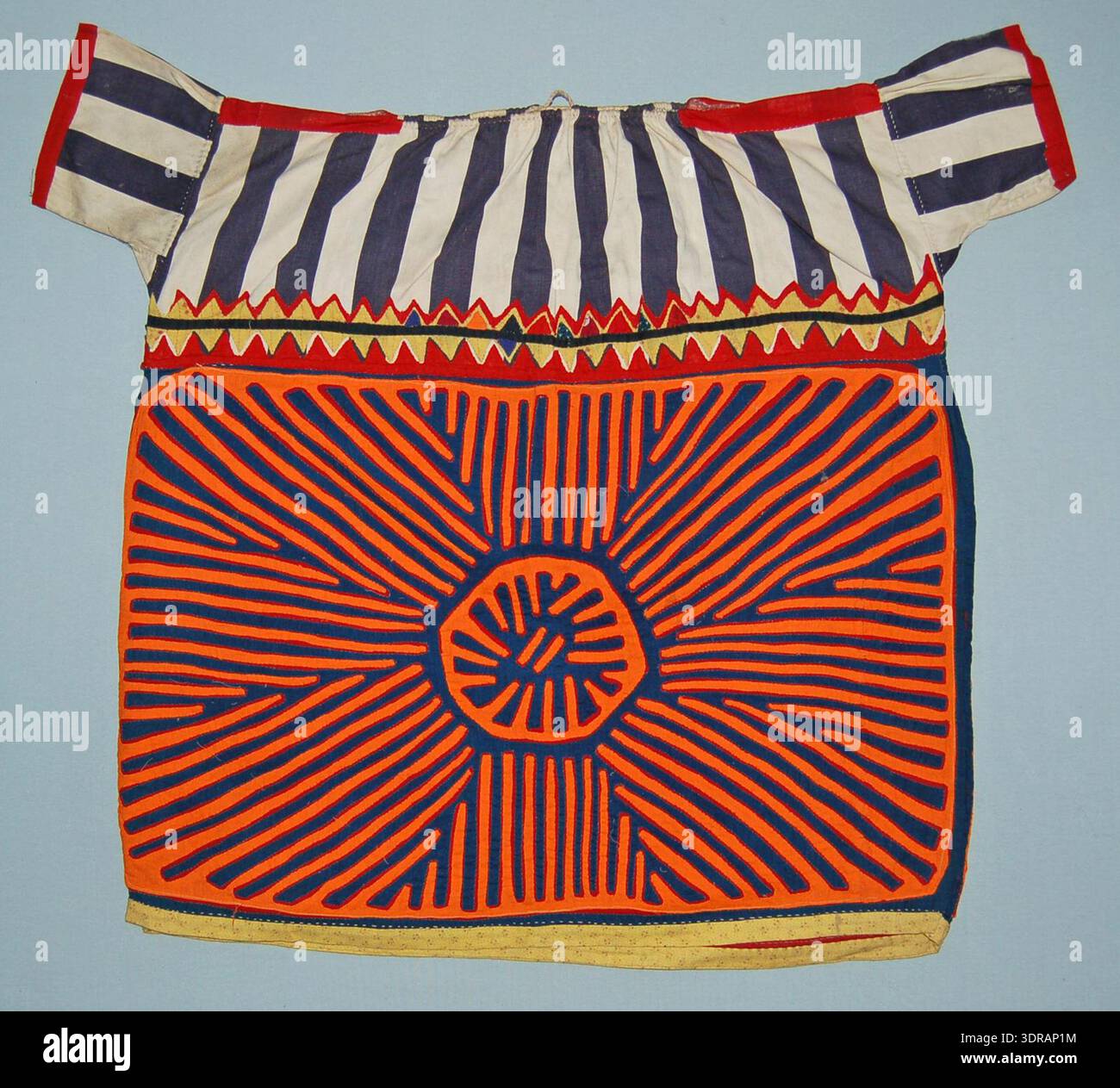Questa blusa mola dei primi del XX secolo è realizzata in cotone, con un giogo di cotone blu stampato con strisce bianche, cotone rosso su spalla e polsini e una fascia di triangoli rossi e patchwork applicati lungo il bordo inferiore. Il supporto del corpo è in cotone rosso, con applicazioni in arancione e blu che raffigurano un design circolare simile al sole. Il cotone giallo completa l'orlo inferiore, mostrando la tradizionale costruzione tessile, tecniche di applicazione indigene, motivi geometrici e pratiche di abbigliamento artigianali. Foto Stock