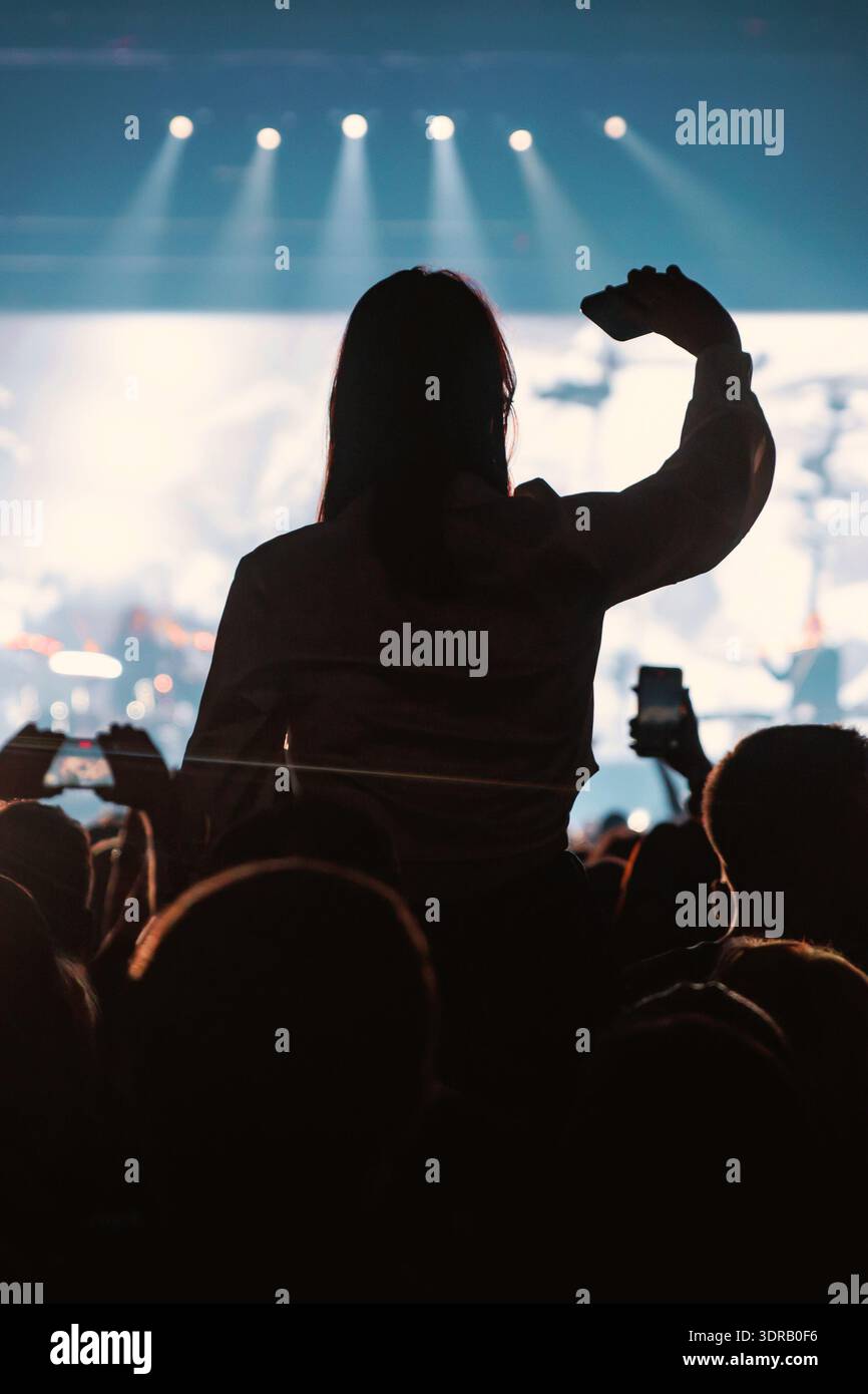 Una persona sagomata si siede sopra la folla per ottenere una visione migliore e registrare un video del palco. Lo sfondo presenta una liscia bianca e blu Foto Stock
