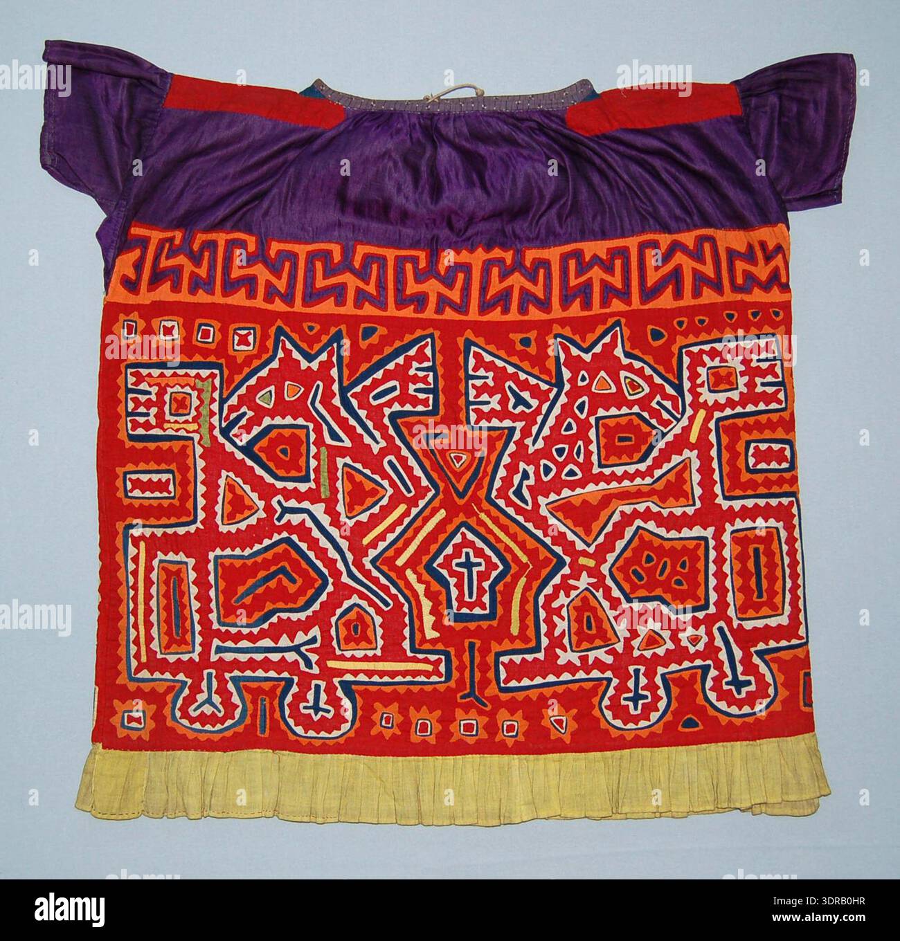 Questa blusa mola dell'inizio del XX secolo è realizzata in cotone con un giogo di rasatello viola, cotone rosso sulle spalle e cotone viola stampato con fiori bianchi cuciti intorno al collo. Una fascia di spirali rosse, gialle e arancioni è applicata intorno al collo. Il supporto del corpo è in cotone blu, con disegni appliqué in rosso, arancione, giallo e bianco, probabilmente raffiguranti un lupo e un volant giallo cucito intorno all'orlo. Illustra le tecniche tessili tradizionali Kuna, l'arte dell'applicazione manuale, la fantasia geometrica e rappresentativa e il design di abbigliamento indigeno. Foto Stock