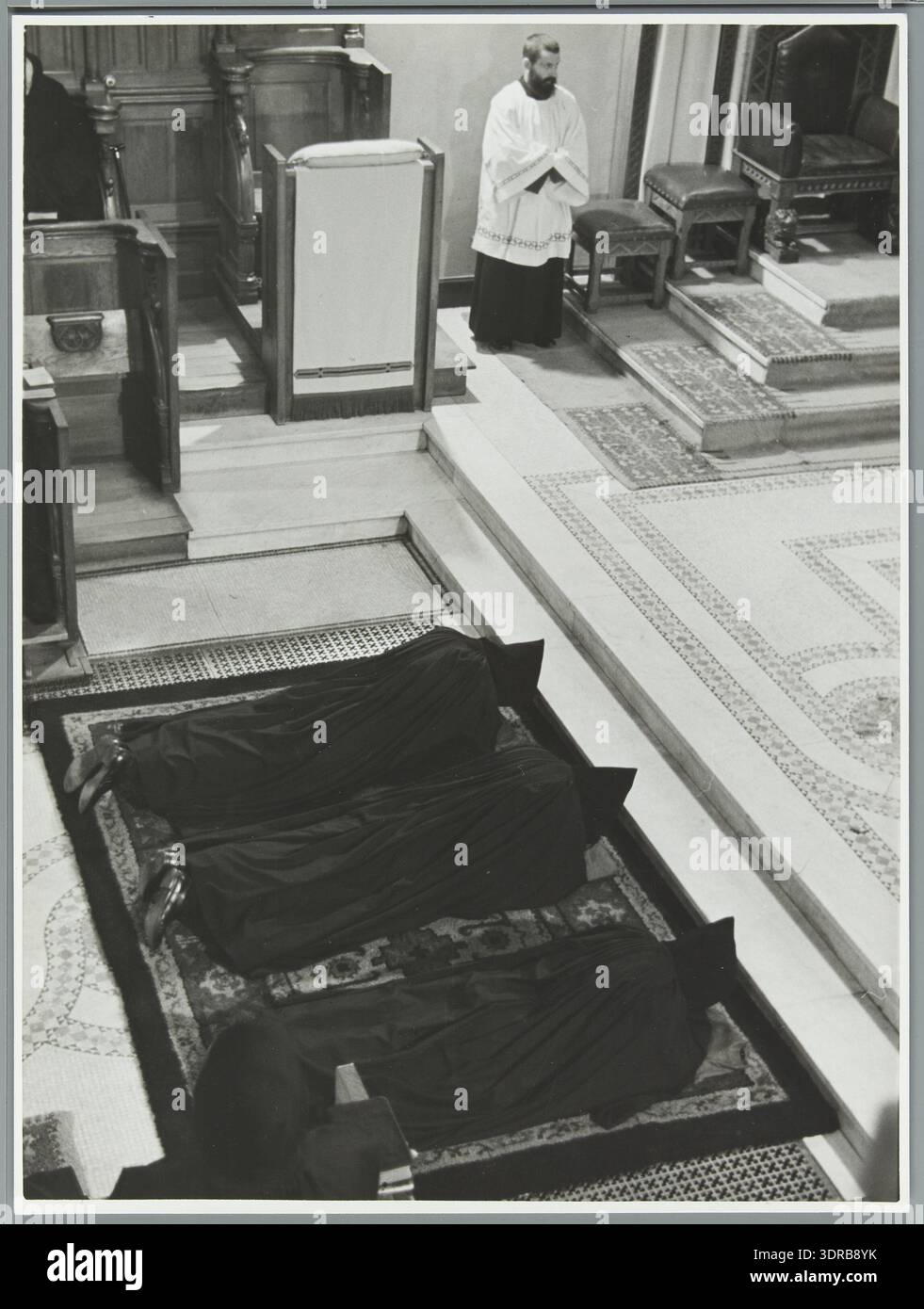 Una fotografia del 1938 dell'abbazia benedettina di St. Andries vicino a Bruges mostra tre monaci stesi su un tappeto sul pavimento, con cappe identiche, durante la cerimonia del "bacio della Pace" dopo aver preso i voti. L'immagine documenta i rituali monastici, la pratica cerimoniale cattolica, le tradizioni benedettine, la messa in scena visiva, la postura liturgica, osservanza religiosa, vita religiosa comune e cultura monastica europea. Foto Stock