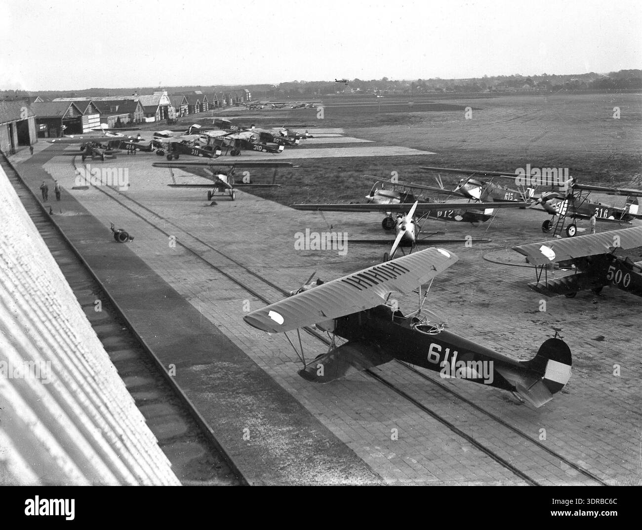 Questa fotografia, scattata da Jan Stevens tra il 1929 e il 1935, mostra la base dell'aeronautica olandese a Soesterberg. Vari tipi di biplani sono parcheggiati davanti agli hangar, compresi gli aerei con numeri di serie 300 e 600. L'immagine documenta l'aviazione militare olandese, i tipi di aerei, le operazioni di hangar, la progettazione di biplani, le infrastrutture dell'aeronautica dell'inizio del XX secolo e il layout della base militare. Foto Stock