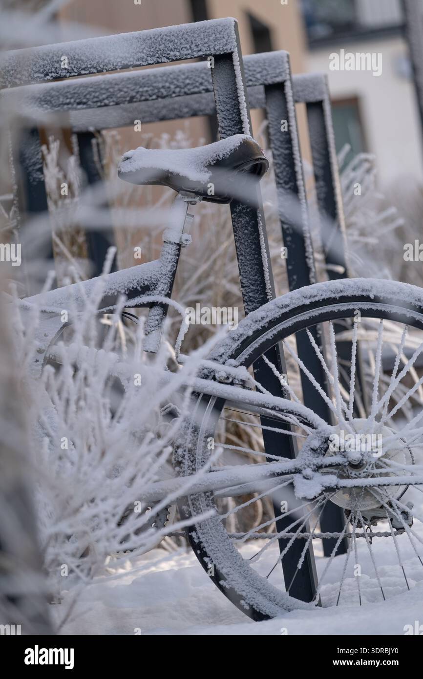 Primo piano di una bicicletta ricoperta di ghiaccio e neve, parcheggiata vicino a una cremagliera metallica in un ambiente urbano invernale. Dettagli ghiacciati su ruota, sella e telaio c Foto Stock
