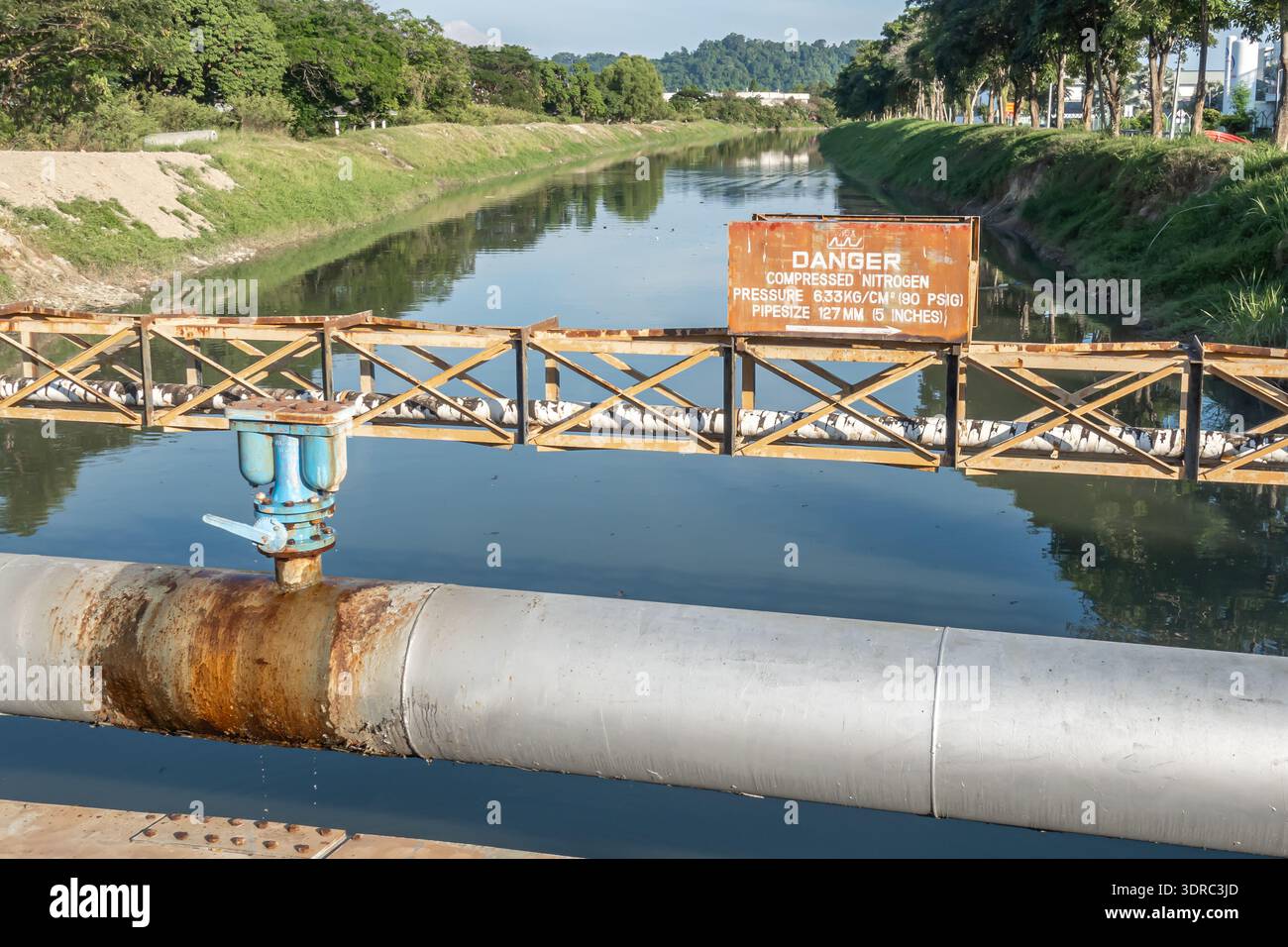 attraversamento di condotte industriali su un canale di drenaggio, un cartello di avvertimento indica che il tubo trasporta azoto compresso. Penang SEZ, Malesia Foto Stock