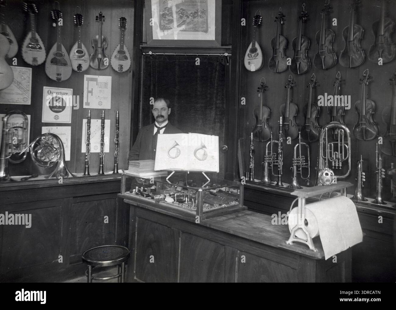Fotografia del 1916 che mostra l'interno del negozio di musica generale 'van Oorbeek & Co' a Keizersgracht 508, angolo di Leidsestraat, Amsterdam. Il negozio espone strumenti musicali, spartiti e accessori. Il proprietario si trova dietro il bancone, interagendo con il personale o i clienti. L'immagine documenta il commercio al dettaglio di musica all'inizio del XX secolo, la sistemazione degli interni e le pratiche di merchandising nei Paesi Bassi. Foto Stock