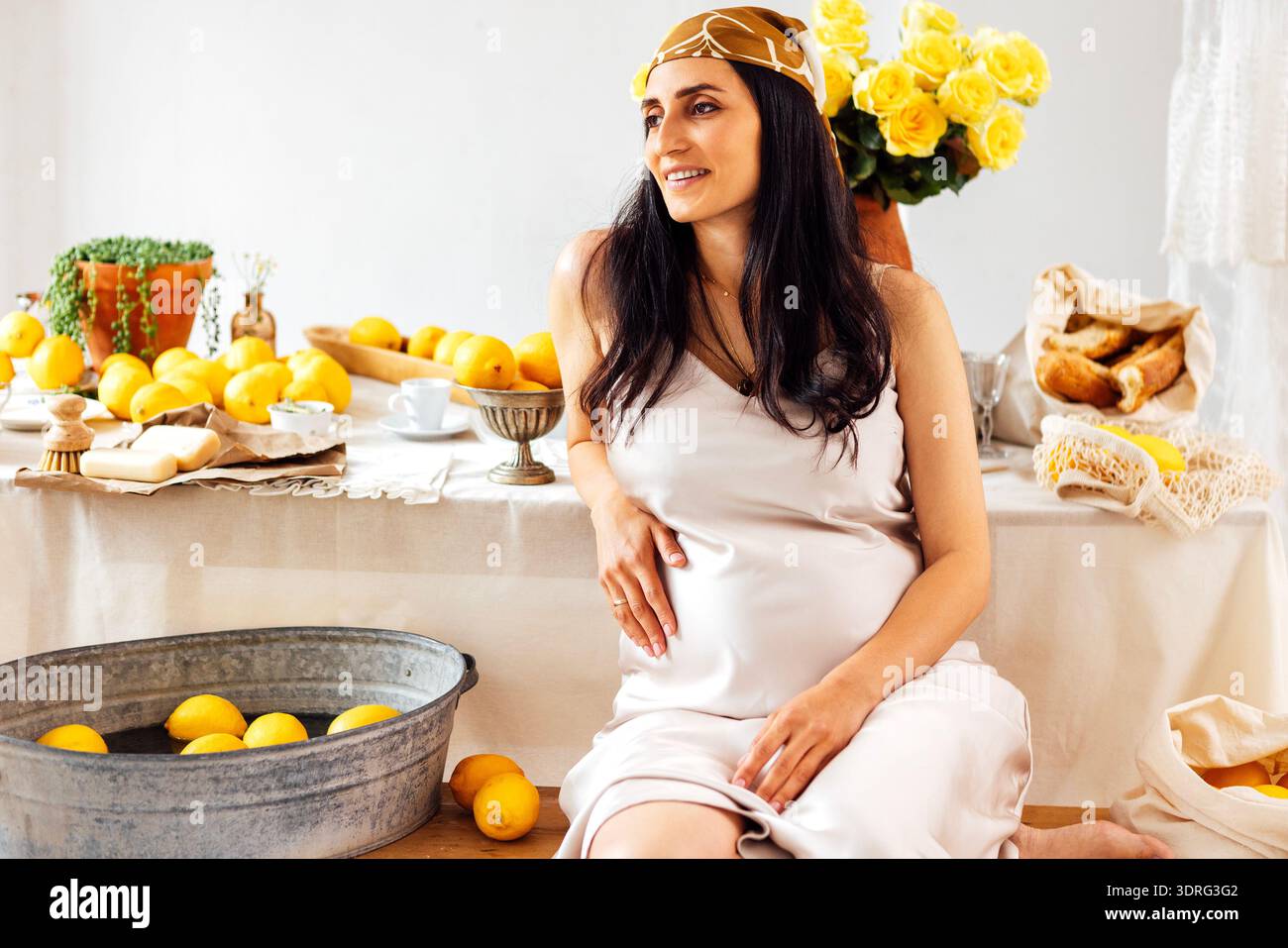 Una donna incinta in un abito di raso siede accanto al tavolo con limoni, utensili da cucina e pane. Lady sorride e tocca delicatamente il suo essere Foto Stock