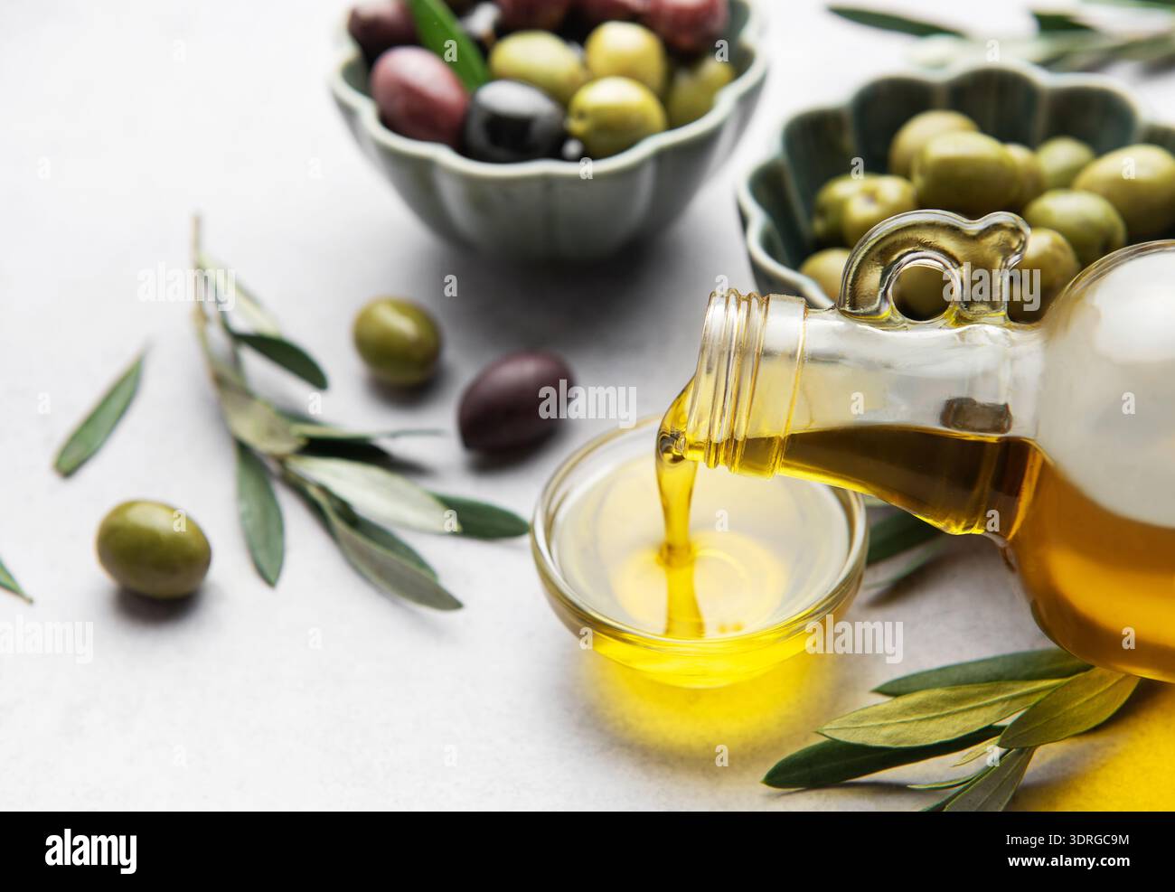 Versare olio d'oliva vergine in una piccola ciotola con olive e rami d'oliva Foto Stock
