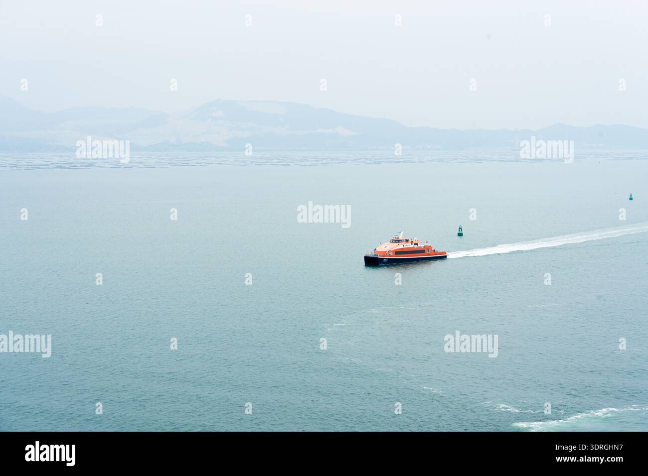Traghetto arancione ad alta velocità che attraversa il mare calmo con colline lontane sotto il cielo coperto Foto Stock