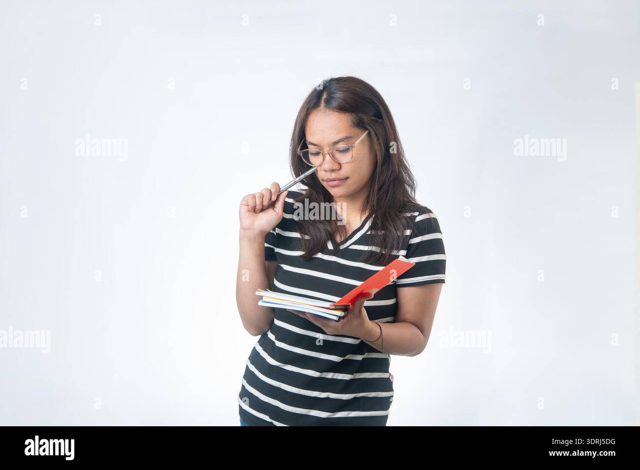 Studente indonesiano concentrato in occhiali che tiene la penna alla fronte mentre legge appunti. Concetto per il duro lavoro, la preparazione degli esami e l'apprendimento mirato. Foto Stock