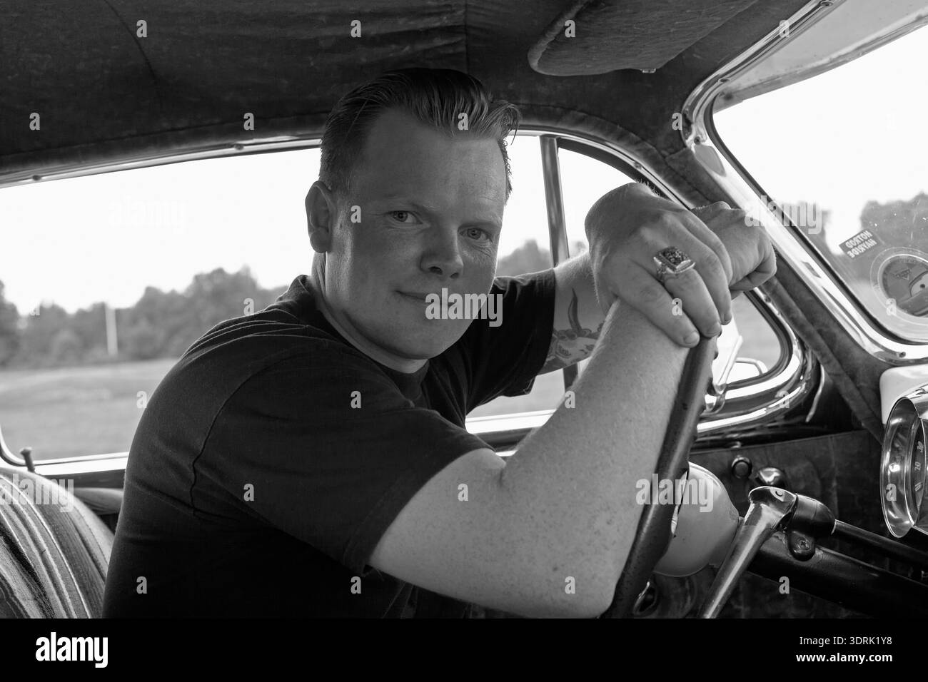 Foto in bianco e nero di un uomo con un soffio seduto al posto di guida di un'auto d'epoca, guardando direttamente la fotocamera. Foto Stock