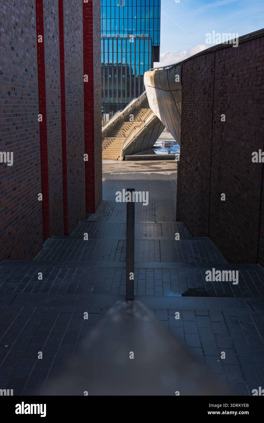 Uno stretto passaggio con pareti di mattoni scuri conduce ad un ponte pedonale scultoreo e ad un alto edificio in vetro. La luce di mezzogiorno proietta ombre nitide, enfatizzando la luminosità Foto Stock