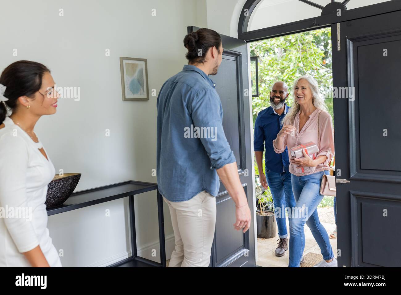 Accogliere gli ospiti a casa, sorridere coppie che salutano gli amici all'ingresso. Ospitalità, amicizia, riunione, arrivo, interazione, Foto Stock