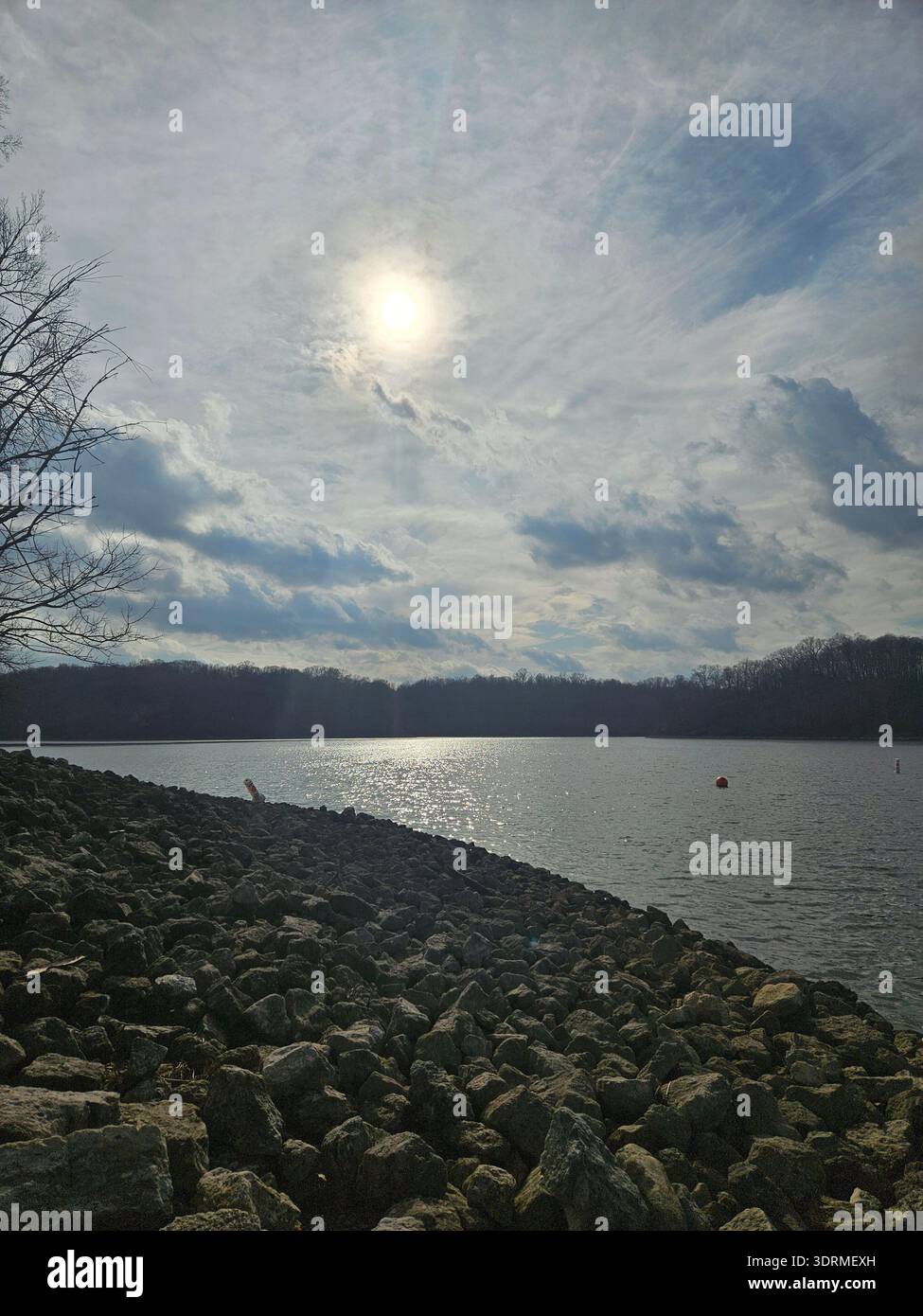 SOLO PER USO EDITORIALE - EAST FORK STATE PARK BETHEL OHIO: ​A fotografia paesaggistica grandangolare del lago William H. Harsha all'East Fork State Park di Bethel, Ohio. La scena presenta una costa rocciosa in primo piano, che conduce alle calme acque soleggiate del bacino idrico. Un luminoso sole pomeridiano pende in un cielo spettacolare pieno di nuvole testurizzate che proiettano un brillante luccichio argentato sulla superficie del lago. L'orizzonte lontano è definito da una fitta linea di alberi della foresta di latifoglie nativa dell'Ohio nel suo inverno dormiente o all'inizio della primavera, catturando la tranquilla bellezza del Midwest wilde - Immagine stock catturata con smartphone