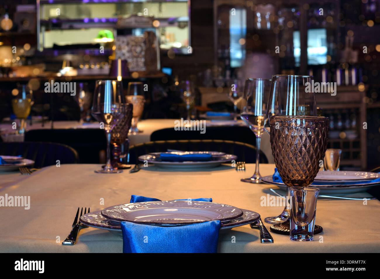 Elegante interno del ristorante con tavoli ben disposti, calici colorati decorativi, bicchieri da vino, piatti bianchi e tovaglioli blu. L'illuminazione calda crea un Foto Stock